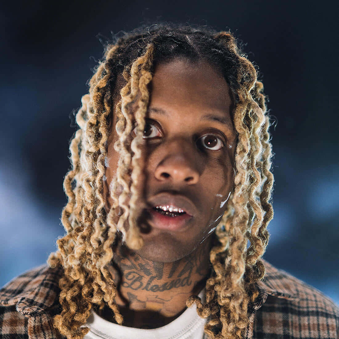 Lil Durk 桌布 壁紙 — 近距離肖像，一名男子留著長長的金色辮子，頸部有刺青，背景是模糊的藍色。
