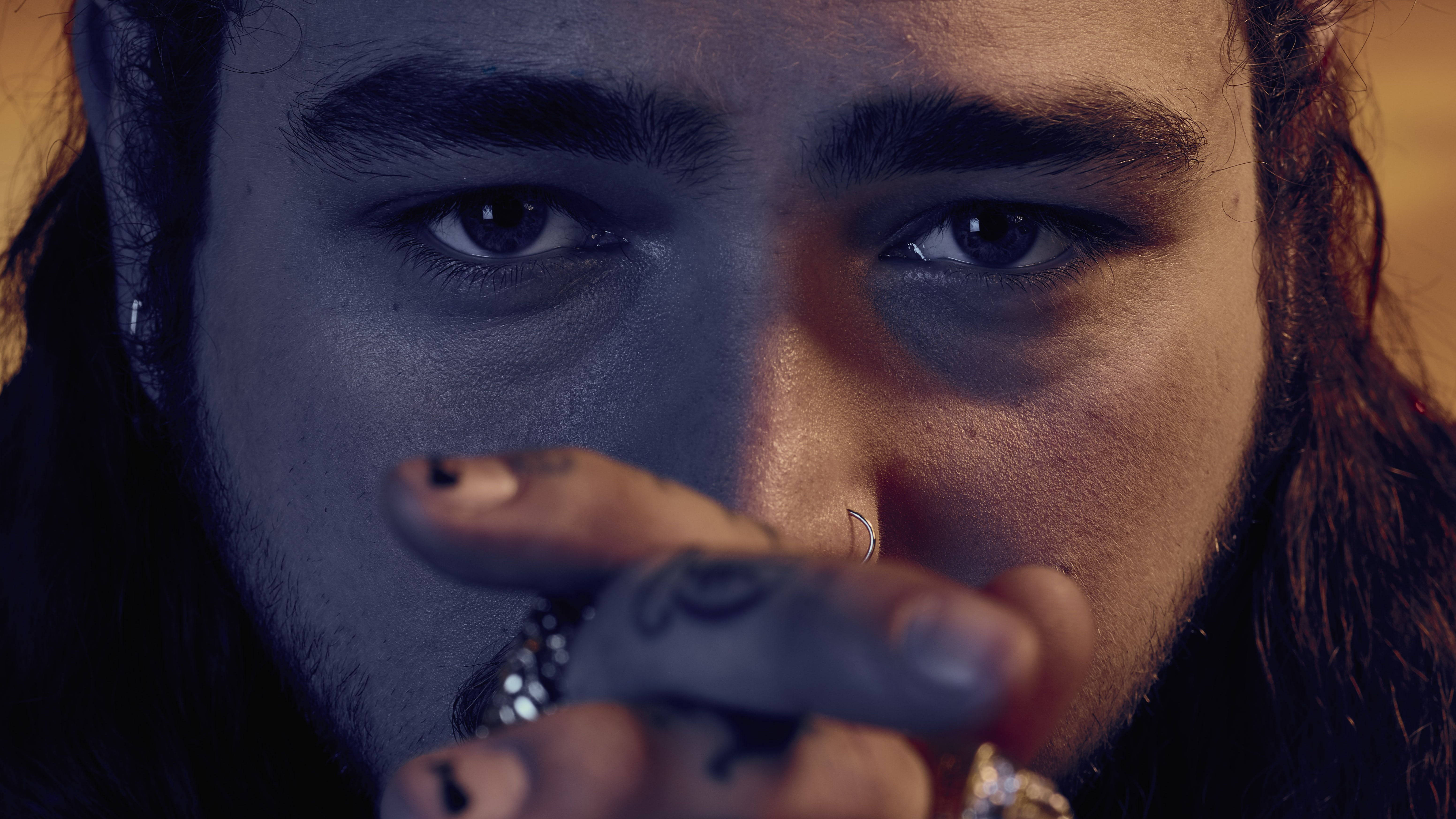 Post Malone 桌布 壁紙 — 近距離肖像，一名眼神銳利、有刺青的男子，在戲劇性的藍色和橙色燈光下，將手放在臉旁。