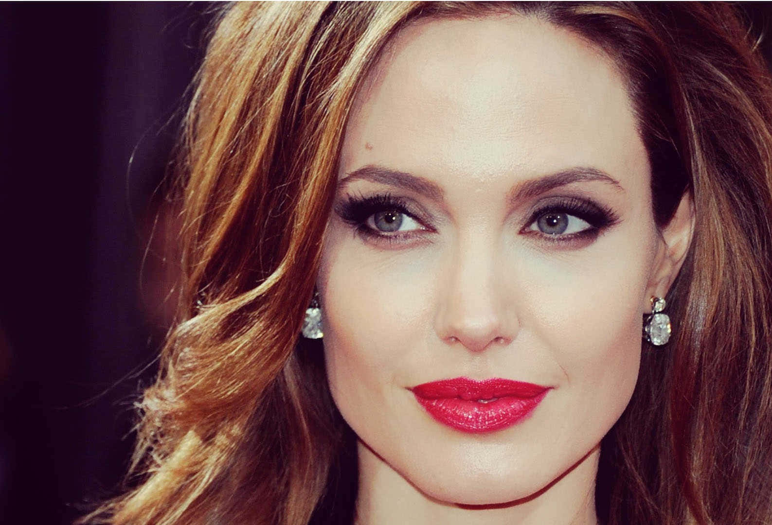 Angelina Jolie 桌布 壁紙 — 近距離肖像照，一位擁有波浪棕色頭髮、大膽紅唇和閃爍鑽石耳環的女性，背景為深色。