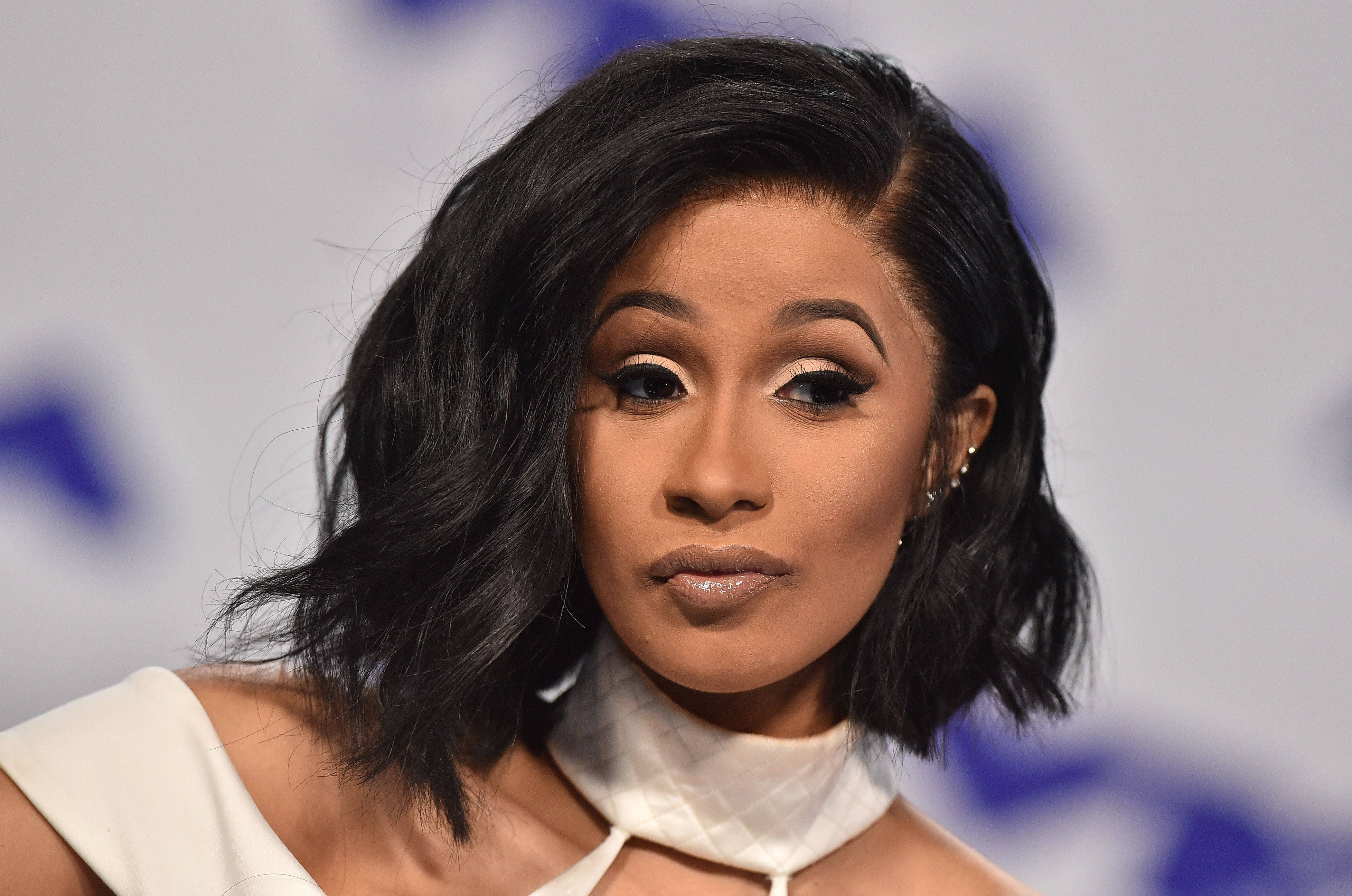 cardi 桌布 壁紙 — 近距離肖像照，一位深色波浪鮑伯頭、妝容大膽的女性穿著白色露肩上衣。