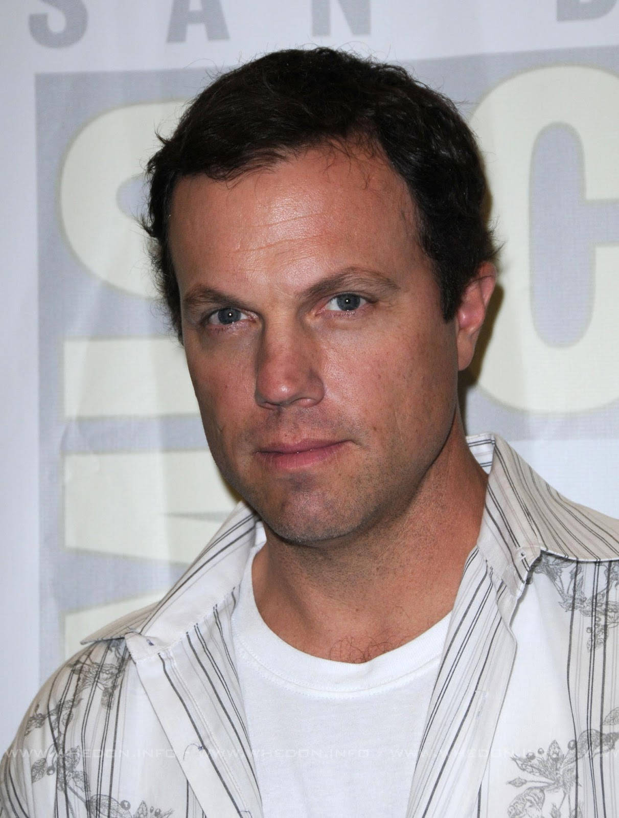 adam baldwin 桌布 壁紙 — 近距離肖像照，一名深色頭髮、眼神銳利的男子穿著圖案襯衫，背景為會議場合。