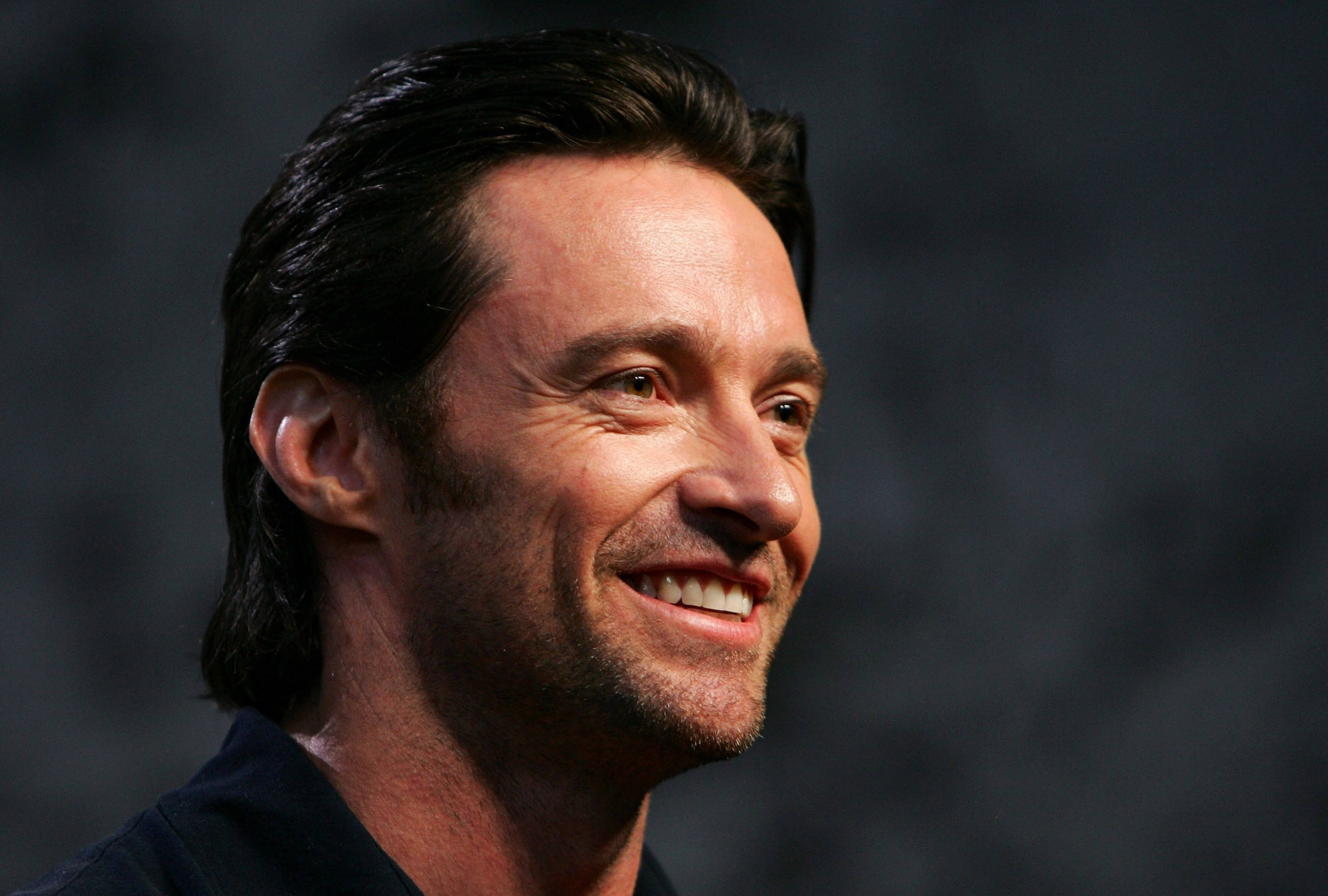 Hugh Jackman 桌布 壁紙 — 近距離肖像照，一名黑髮、鬍渣的男子微笑，背景為模糊灰色。