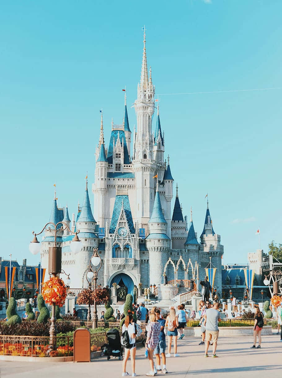 Disney Castle 桌布 壁紙 — 迪士尼灰姑娘城堡晴朗的一天，遊客在晴朗的藍天下圍繞其宏偉的藍白塔樓漫步。