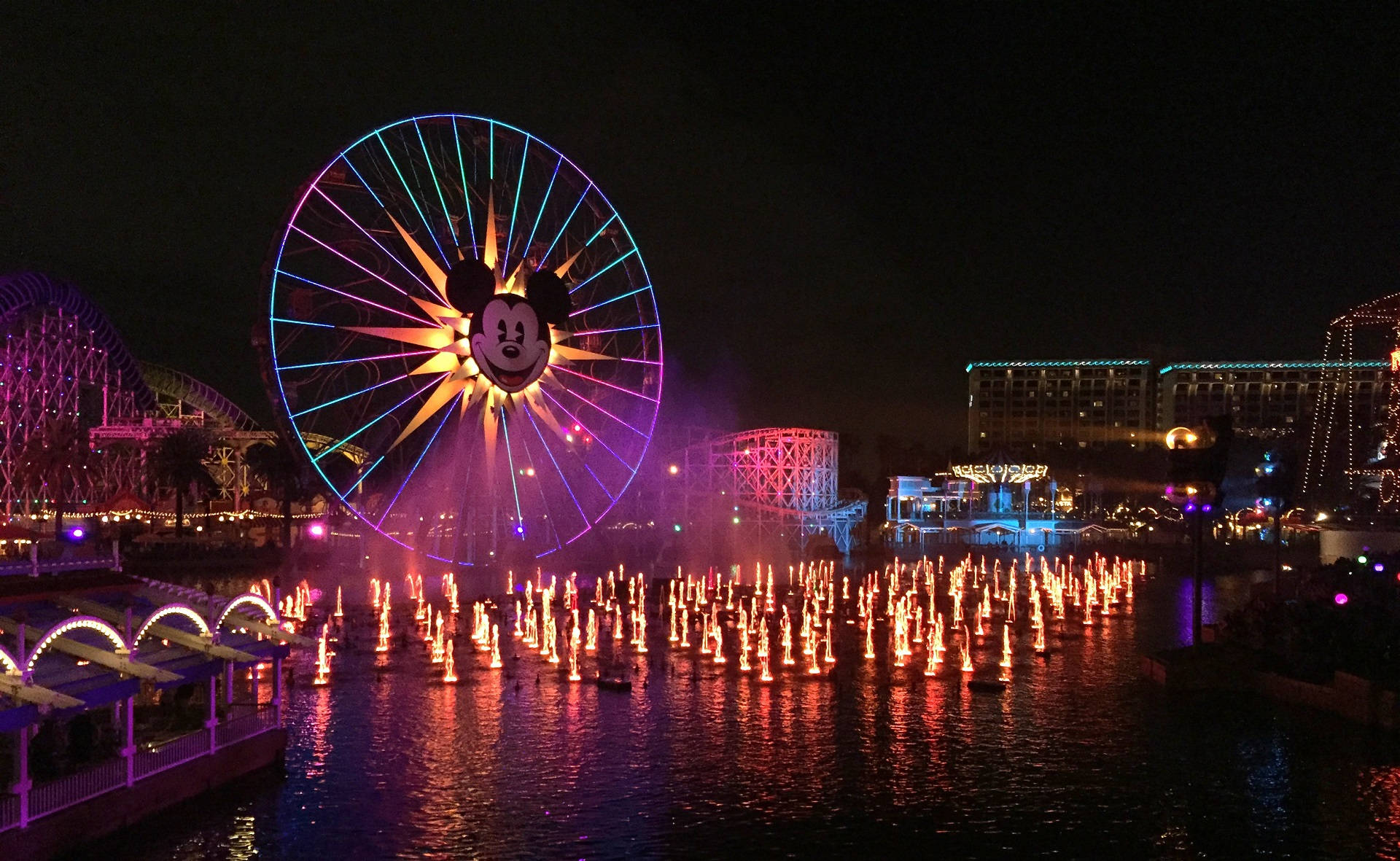 Disneyland Desktop 桌布 壁紙 — 迪士尼的迷人夜景，巨大的米奇老鼠摩天輪在色彩繽紛的噴泉上方閃耀，噴泉將彩色燈光反射在水面上。