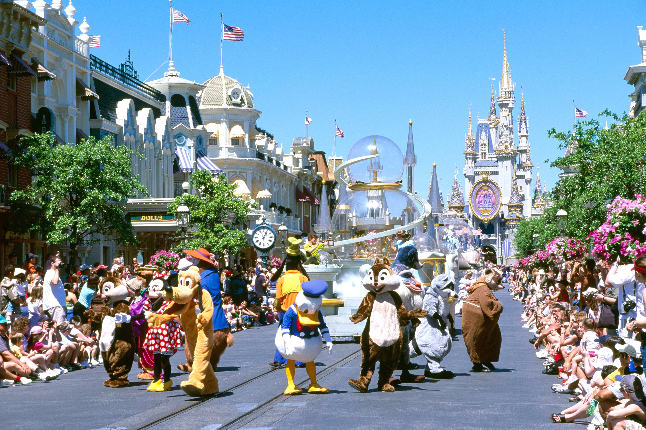 Disneyworld 桌布 壁紙 — 迪士尼遊行角色在陽光明媚的街道上行進，街道兩旁擠滿人群和標誌性的城堡建築。