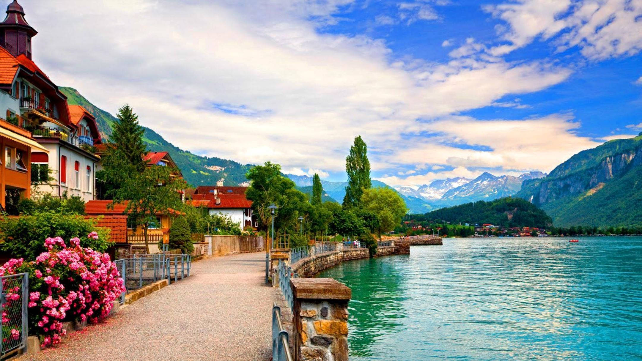 Switzerland 桌布 壁紙 — 迷人的湖邊小徑，兩旁是色彩繽紛的房屋和粉紅色的花朵，頭頂是明亮的藍天和蓬鬆的白雲。