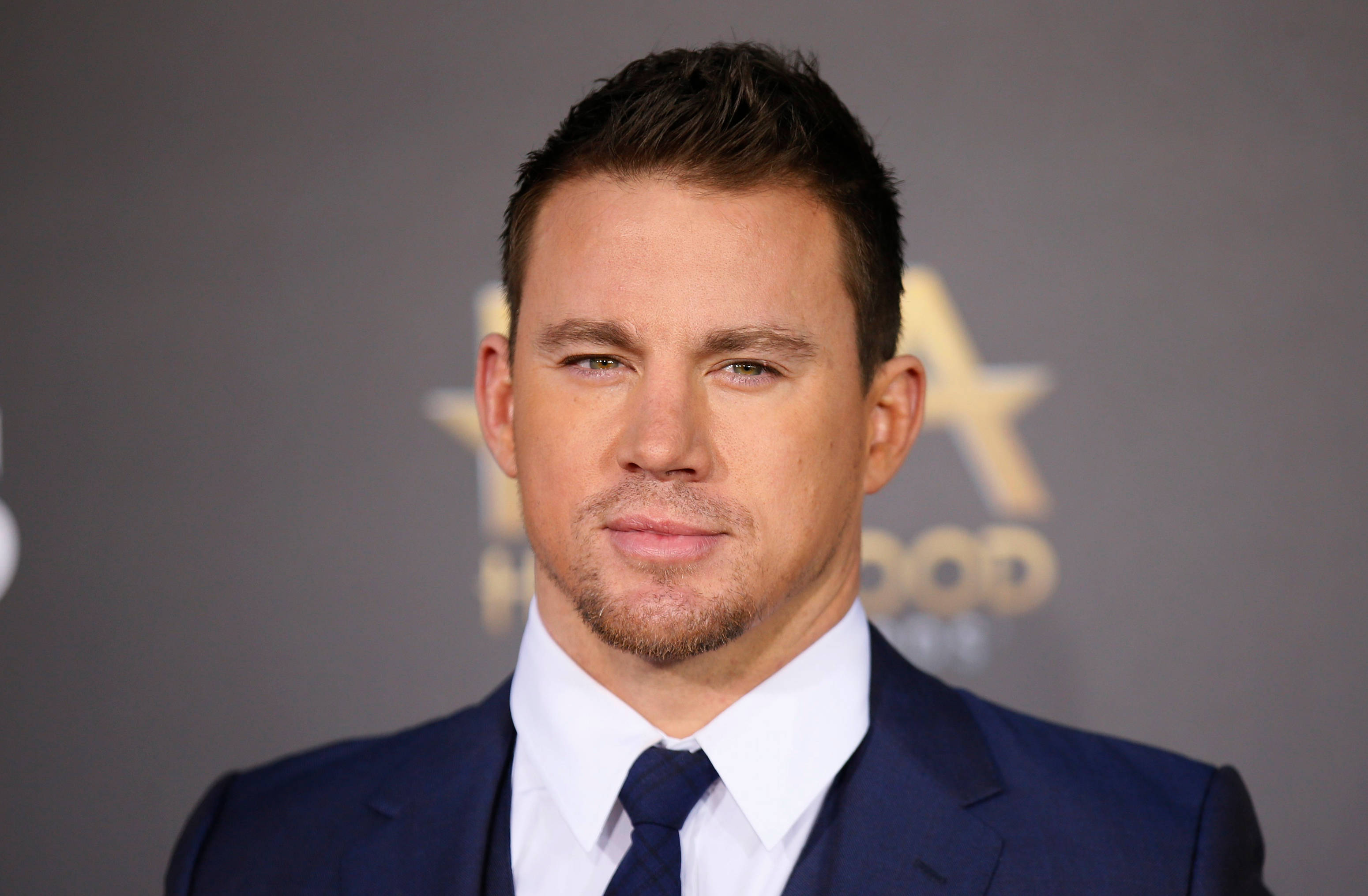 Channing 桌布 壁紙 — 迷人的演員穿著俐落的深藍色西裝和領帶，在帶有金色星星標誌的柔和灰色背景前自信地擺姿勢。