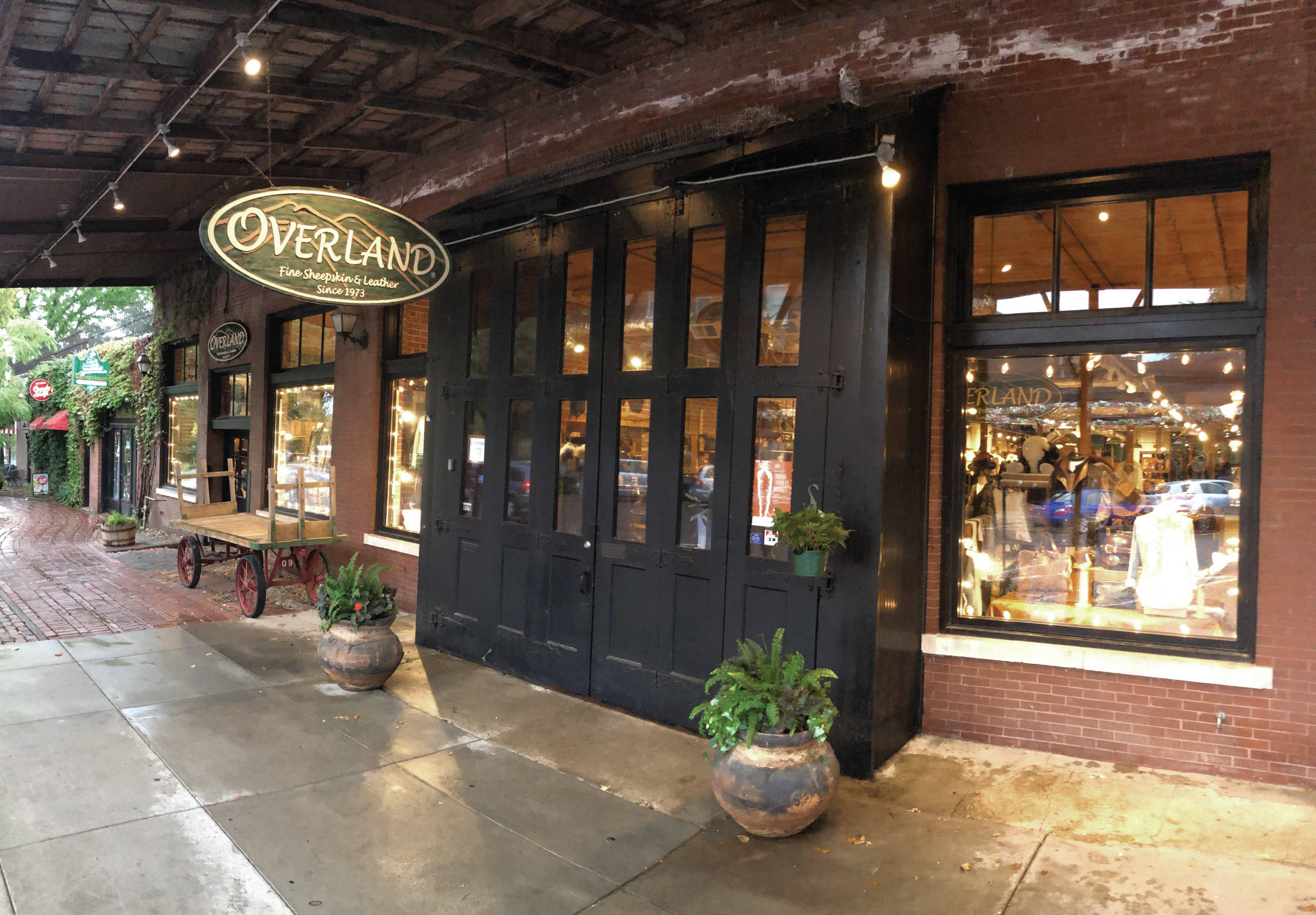 Store 桌布 壁紙 — 迷人的磚砌店面，黑色門窗在潮濕的人行道上，在溫暖的串燈下展示皮革製品。