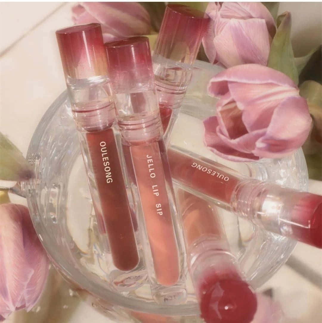 lipstick 桌布 壁紙 — 透明玻璃碗盛裝粉紅色唇彩管，置於柔和的粉紅色鬱金香之間，沐浴在溫暖的粉彩光線中。