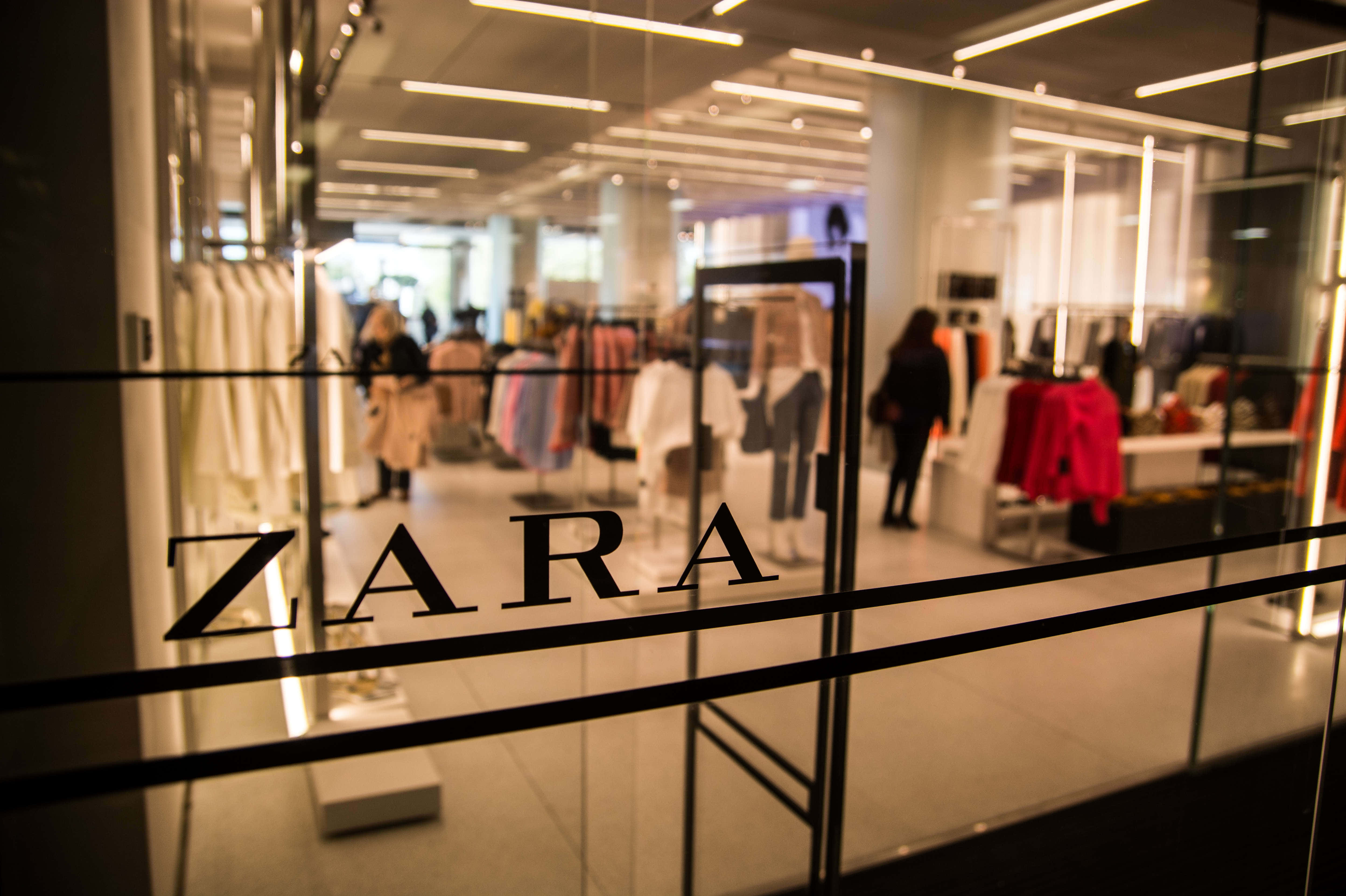 Zara 桌布 壁紙 — 透過Zara商店玻璃門的模糊視野，顯示顧客在明亮天花板燈光下瀏覽色彩繽紛的服裝架。