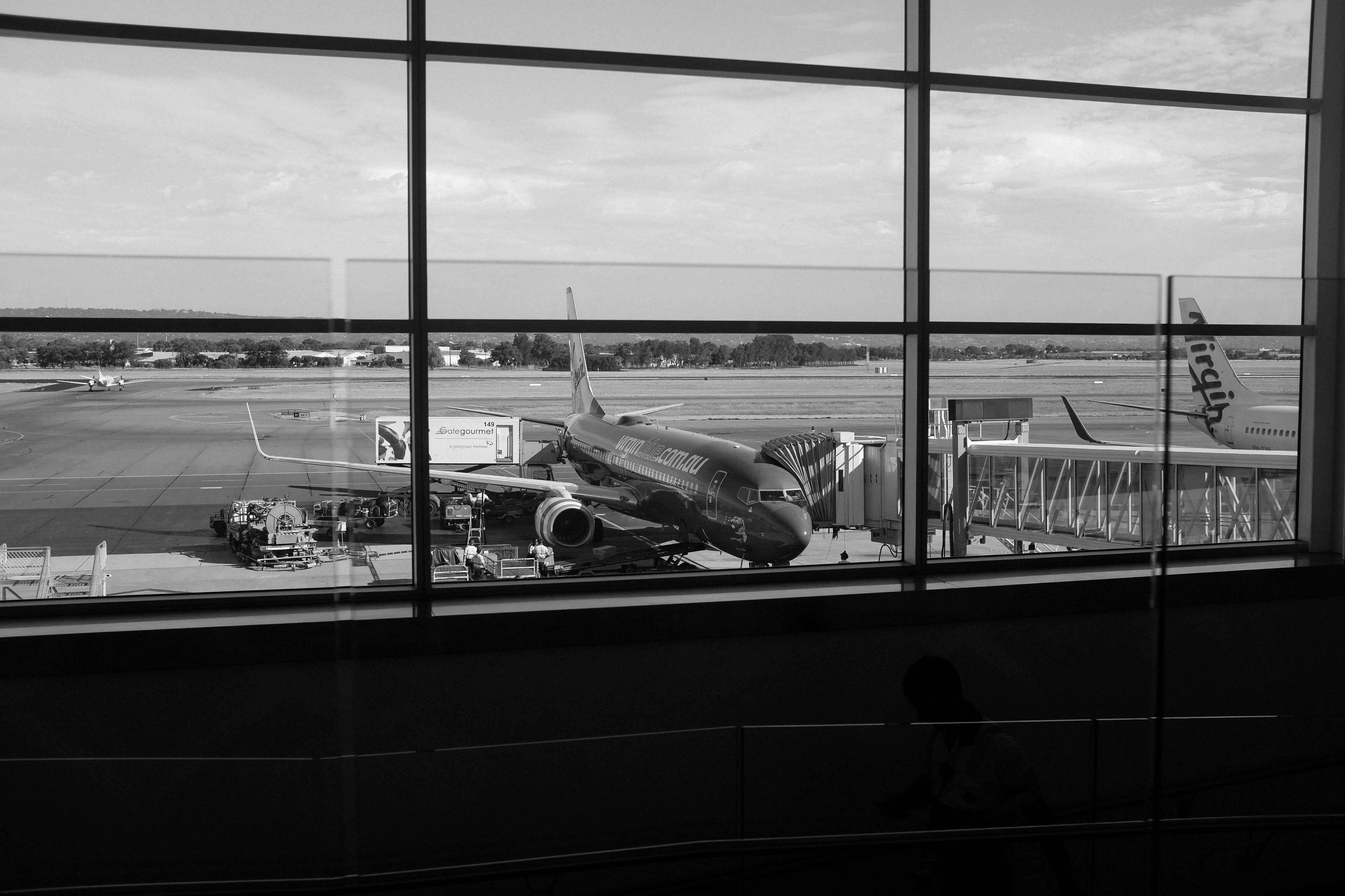 Airport 桌布 壁紙 — 透過大窗戶看到的黑白飛機停泊在機場閘口的景色，前景有乘客剪影。