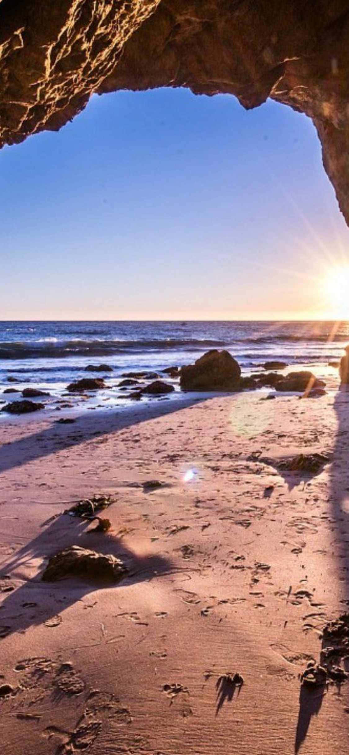 iPhone Xs Malibu 桌布 壁紙 — 透過岩石洞穴開口看到的日落，映照在沙灘上，伴隨著海浪和散落的巨石。