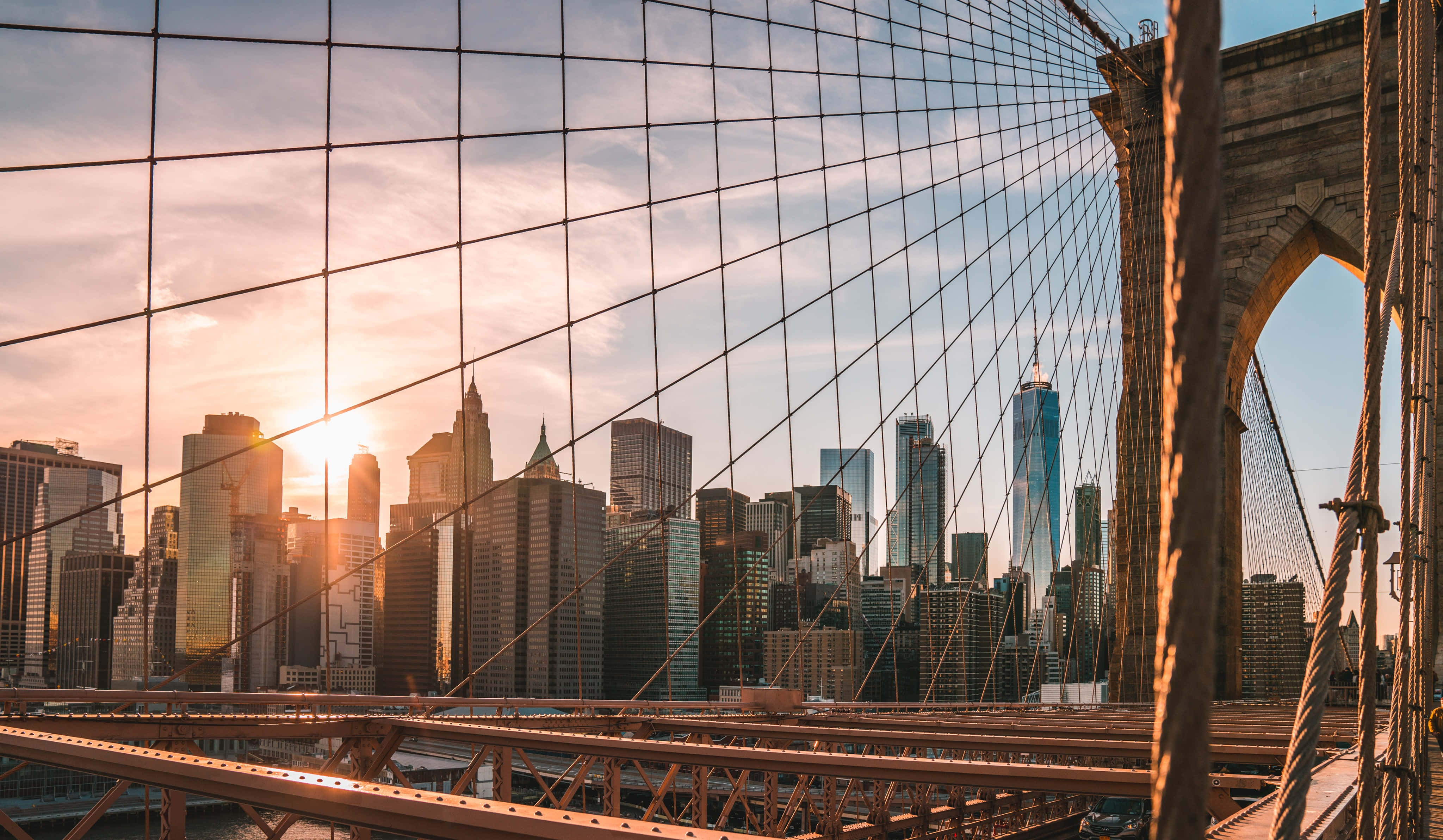 Brooklyn Bridge 桌布 壁紙 — 透過布魯克林大橋複雜的鋼纜，看到紐約市摩天大樓背後的金色夕陽閃耀。