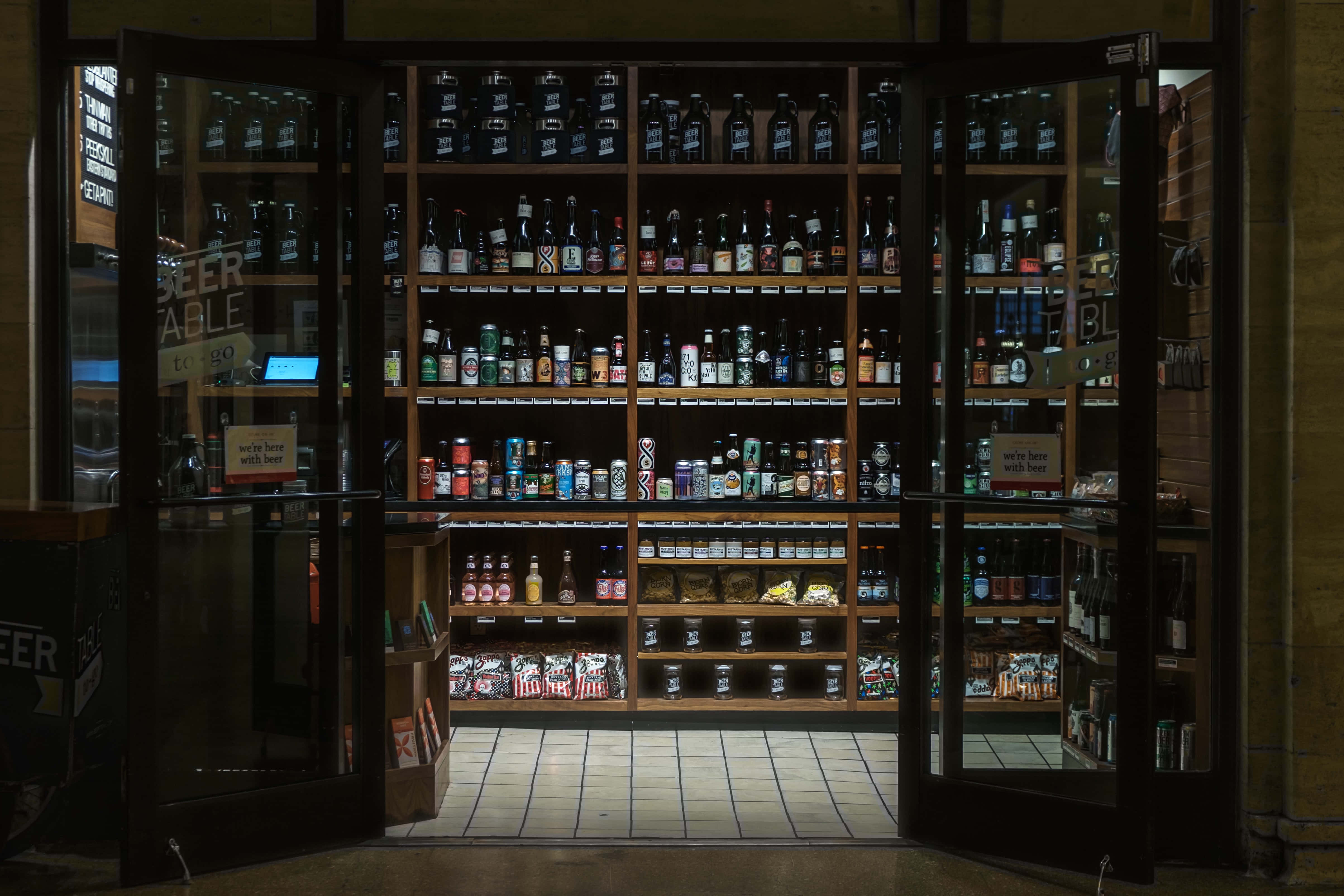 高清 桌布 壁紙 — 透過打開的玻璃門看到的溫暖燈光啤酒商店內部，展示著擺滿數百個瓶子和罐子的貨架。