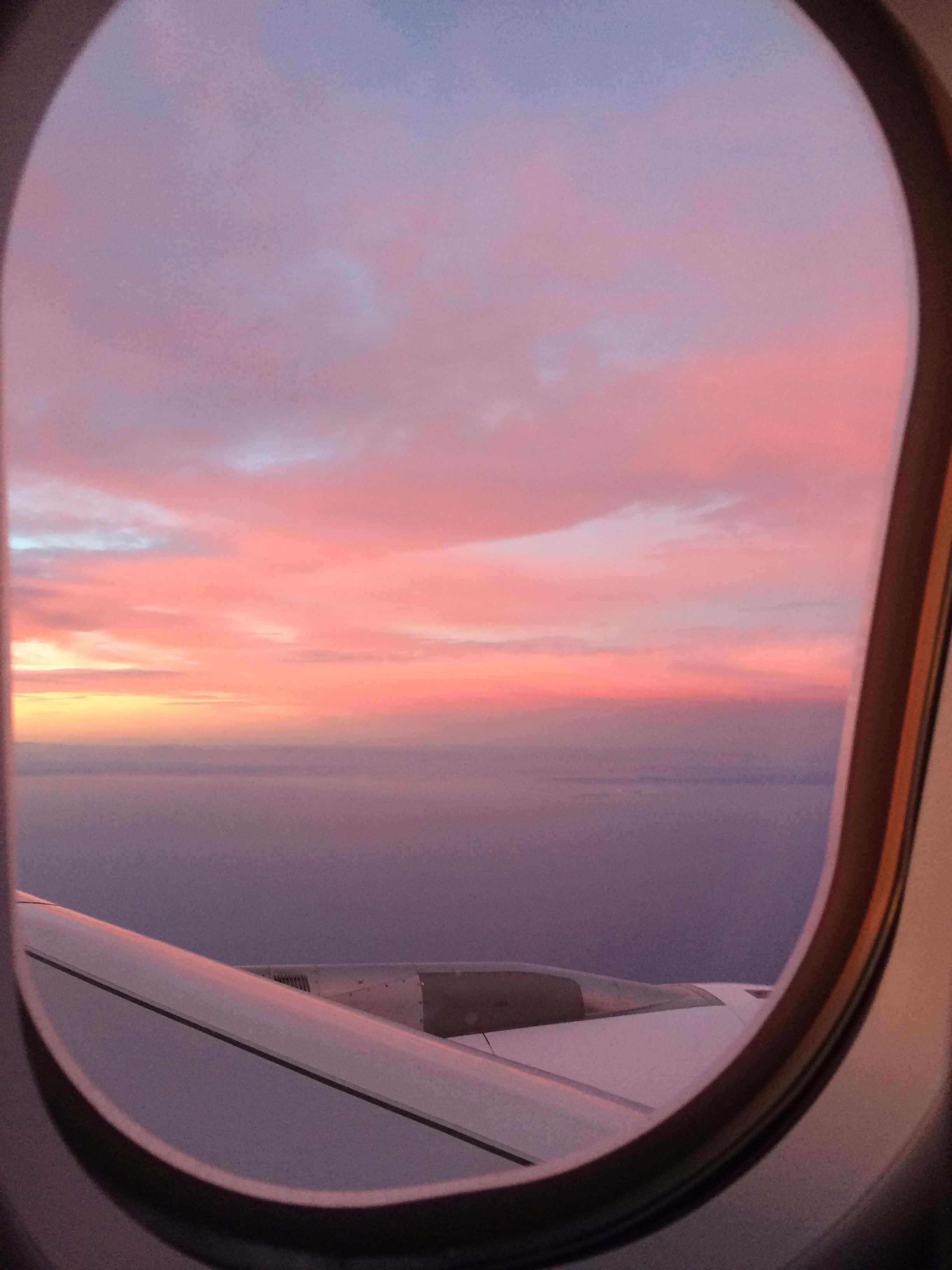 Plane 桌布 壁紙 — 透過橢圓形窗戶看到的飛機機翼，粉橙色日落將雲彩染上色彩，映襯著無盡的海洋地平線。