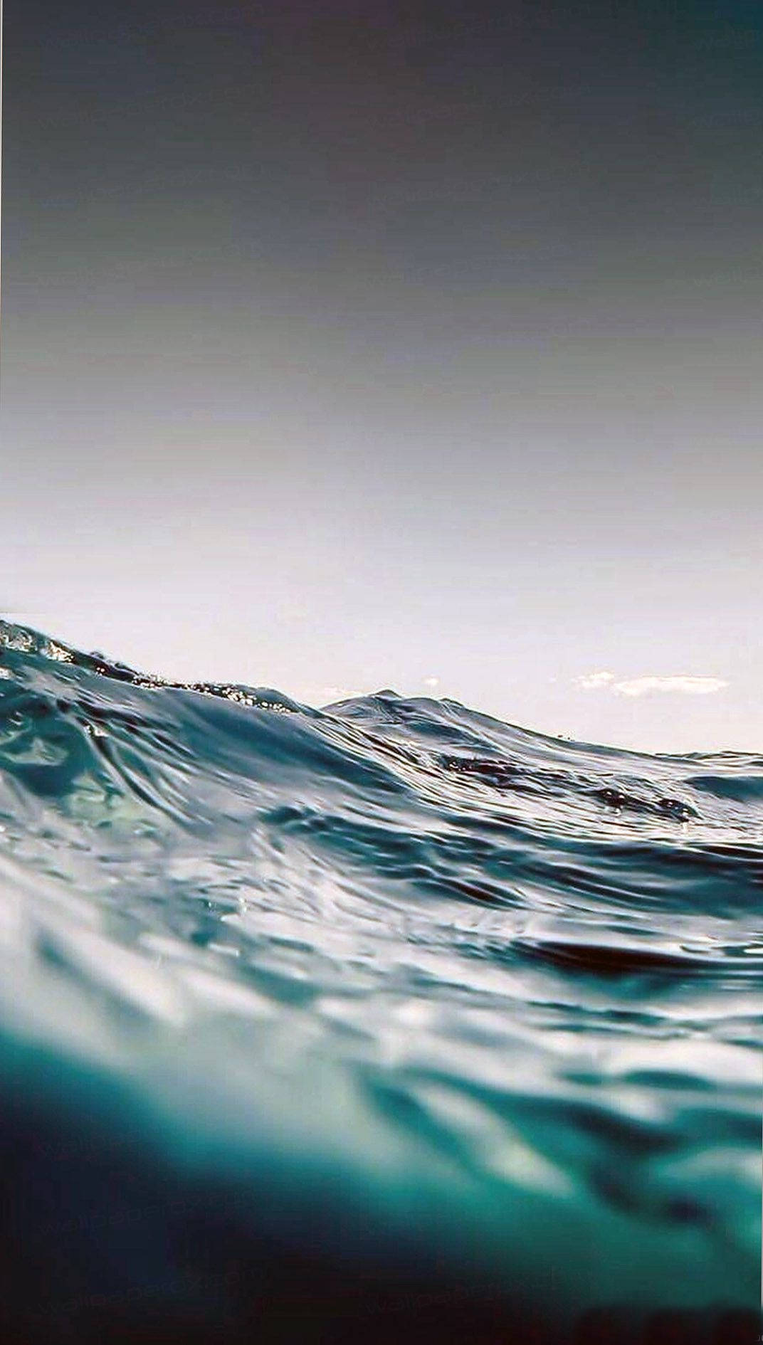 Ocean iPhone 桌布 壁紙 — 透過漣漪的碧綠波浪向上望向柔和灰色天空的寧靜水下景色。