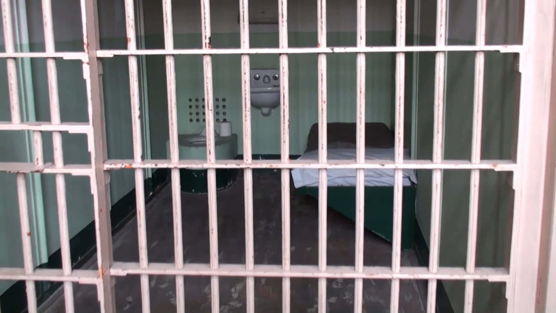 Prison Cell 桌布 壁紙 — 透過生鏽的金屬欄杆看到的陰森監獄牢房，裡面有一張簡單的床和水槽。