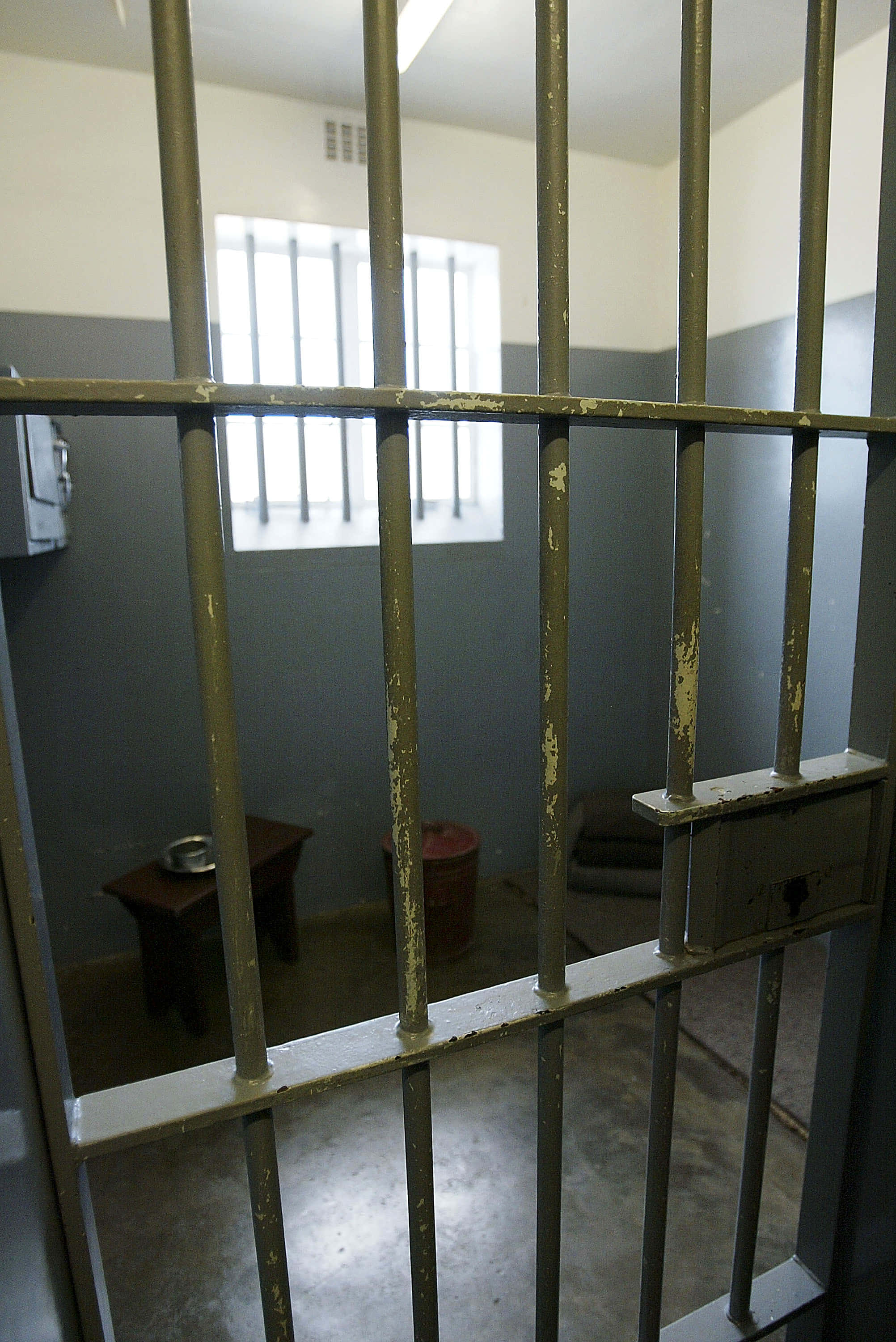 Jail Bars 桌布 壁紙 — 透過生鏽金屬欄杆看到的昏暗監獄牢房，設有小窗戶和基本家具。