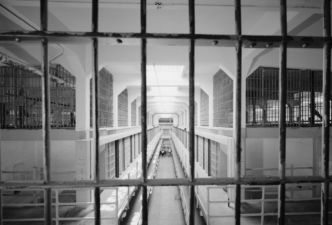 Prison 桌布 壁紙 — 透過監獄鐵欄杆看到的黑白景象，是一條長長的機構走廊，有多層牢房和遠處的人影。