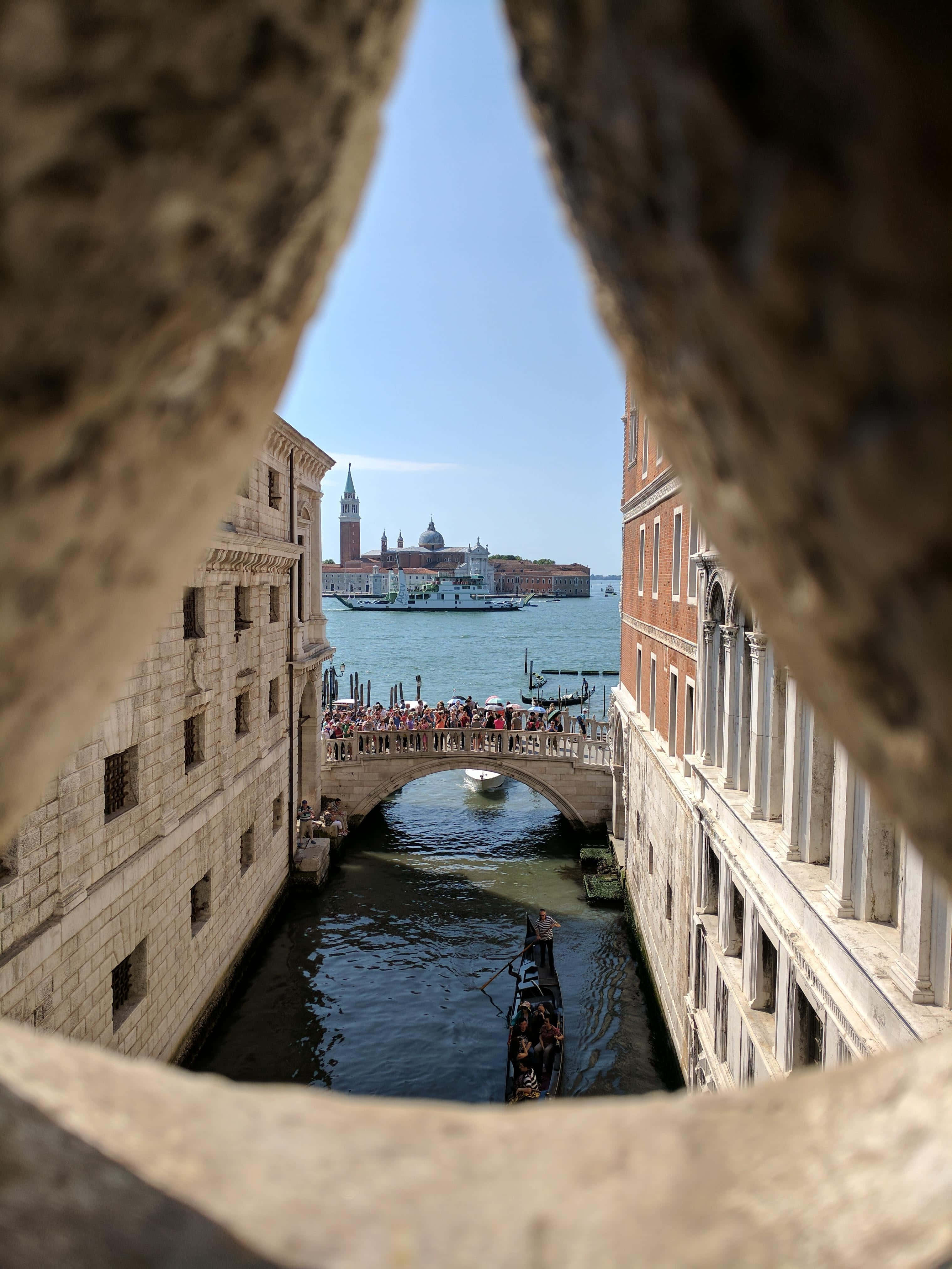 bridge 桌布 壁紙 — 透過石拱門的景色，框住了威尼斯運河，有貢多拉和一座擠滿人群的橋樑，在清澈的藍天下。
