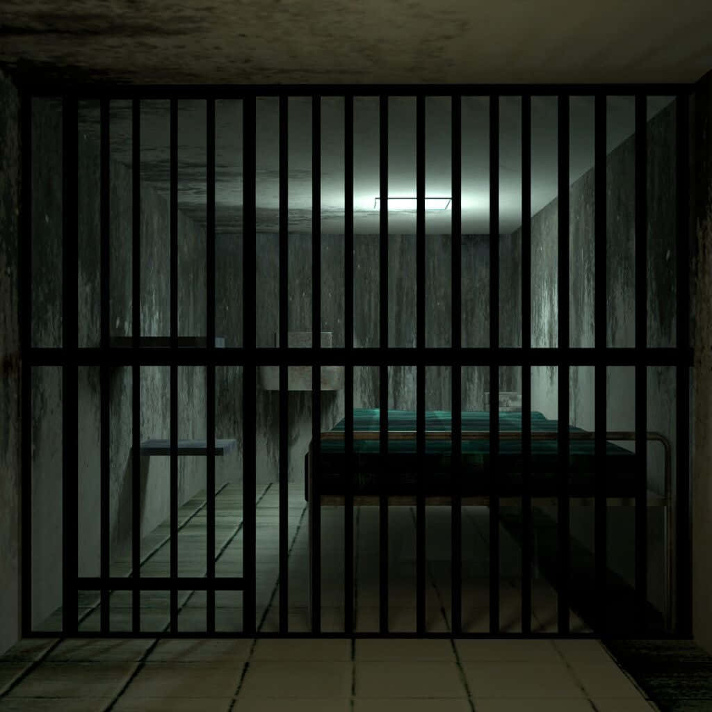 Prison Cell 桌布 壁紙 — 透過金屬欄杆看到的昏暗監獄牢房，有一張單人床和混凝土牆，天花板上閃爍的燈光。
