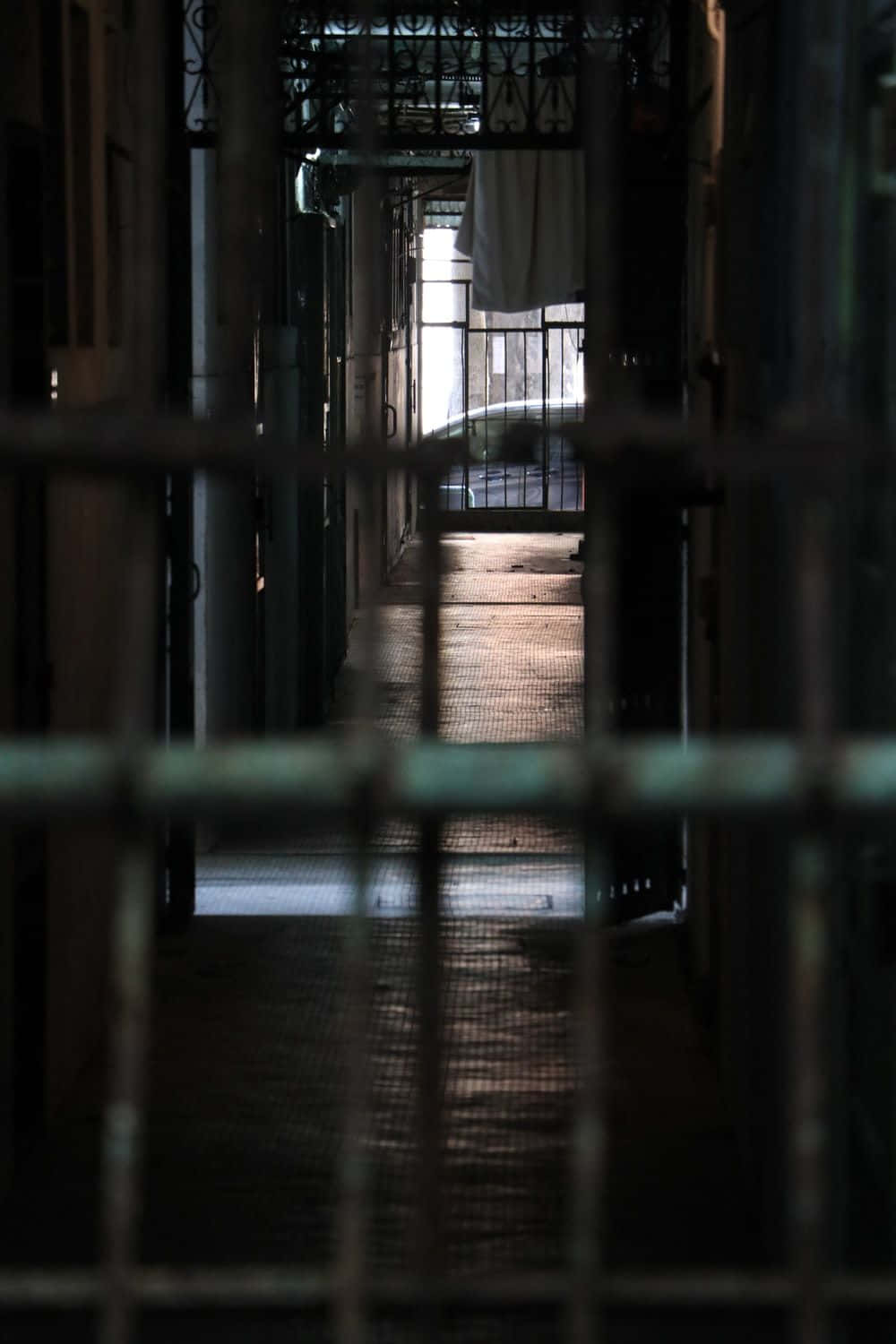 Prison Cell 桌布 壁紙 — 透過金屬欄杆看到的昏暗監獄走廊，遠處掛著一條毛巾，盡頭有光。