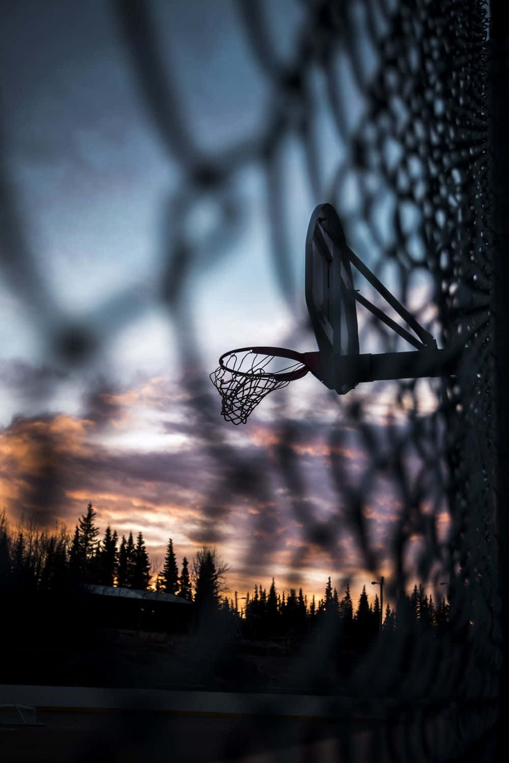 Basketball Aesthetic 桌布 壁紙 — 透過鐵絲網圍欄看到的籃球框，在充滿活力的日落天空下形成剪影。