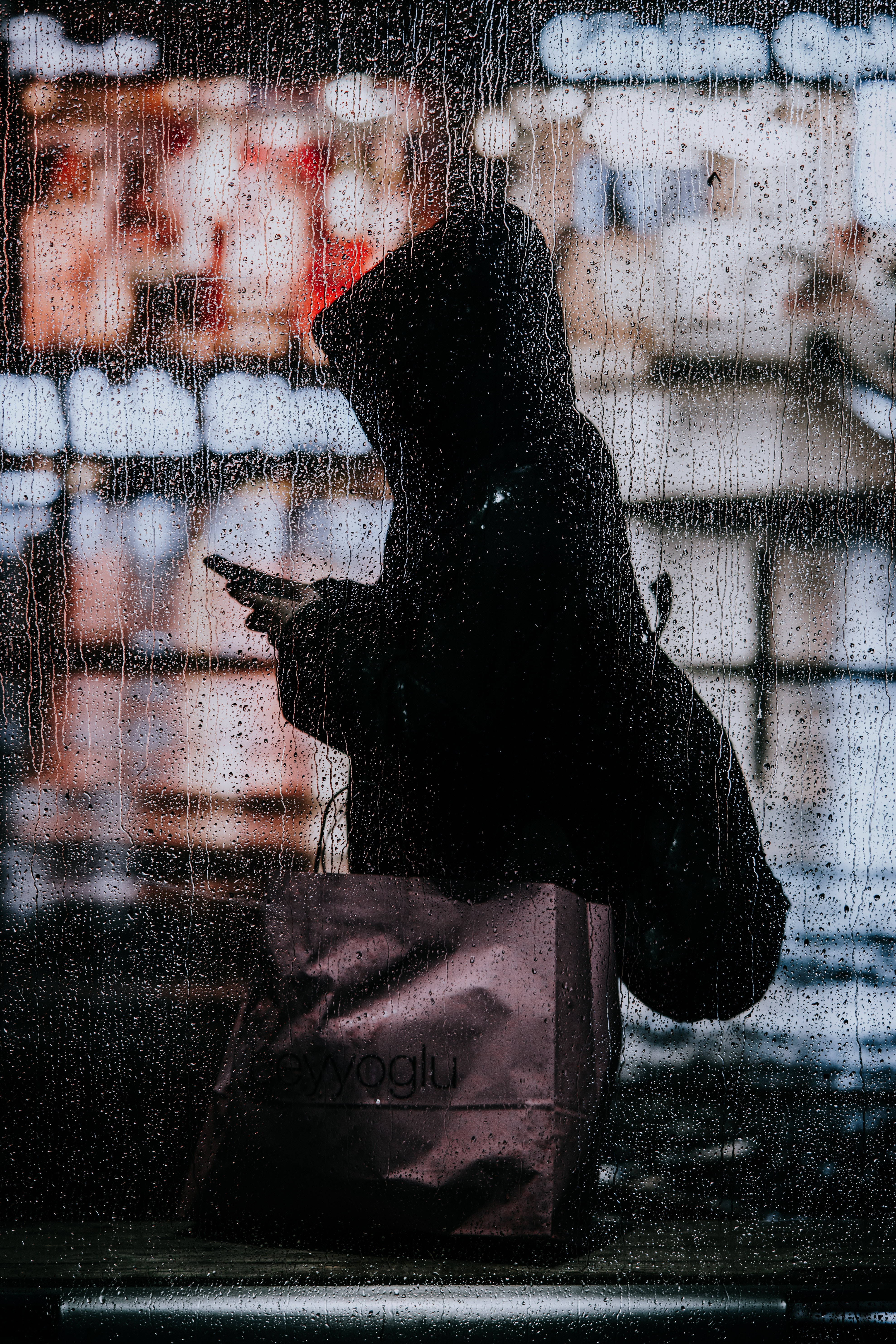 下雨 桌布 壁紙 — 透過雨痕玻璃看到的戴帽人物剪影，手持電話和購物袋，背後是模糊的城市燈光。