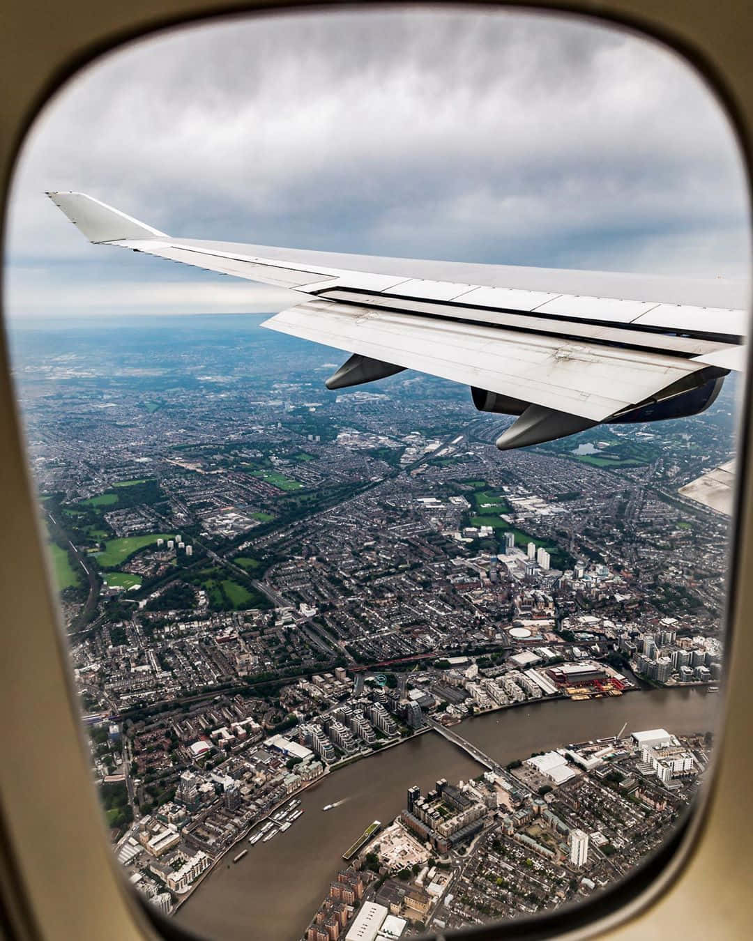 Airplane Window 桌布 壁紙 — 透過飛機窗戶俯瞰，機翼下方是廣闊的城市景觀，河流和橋樑在陰天之下。