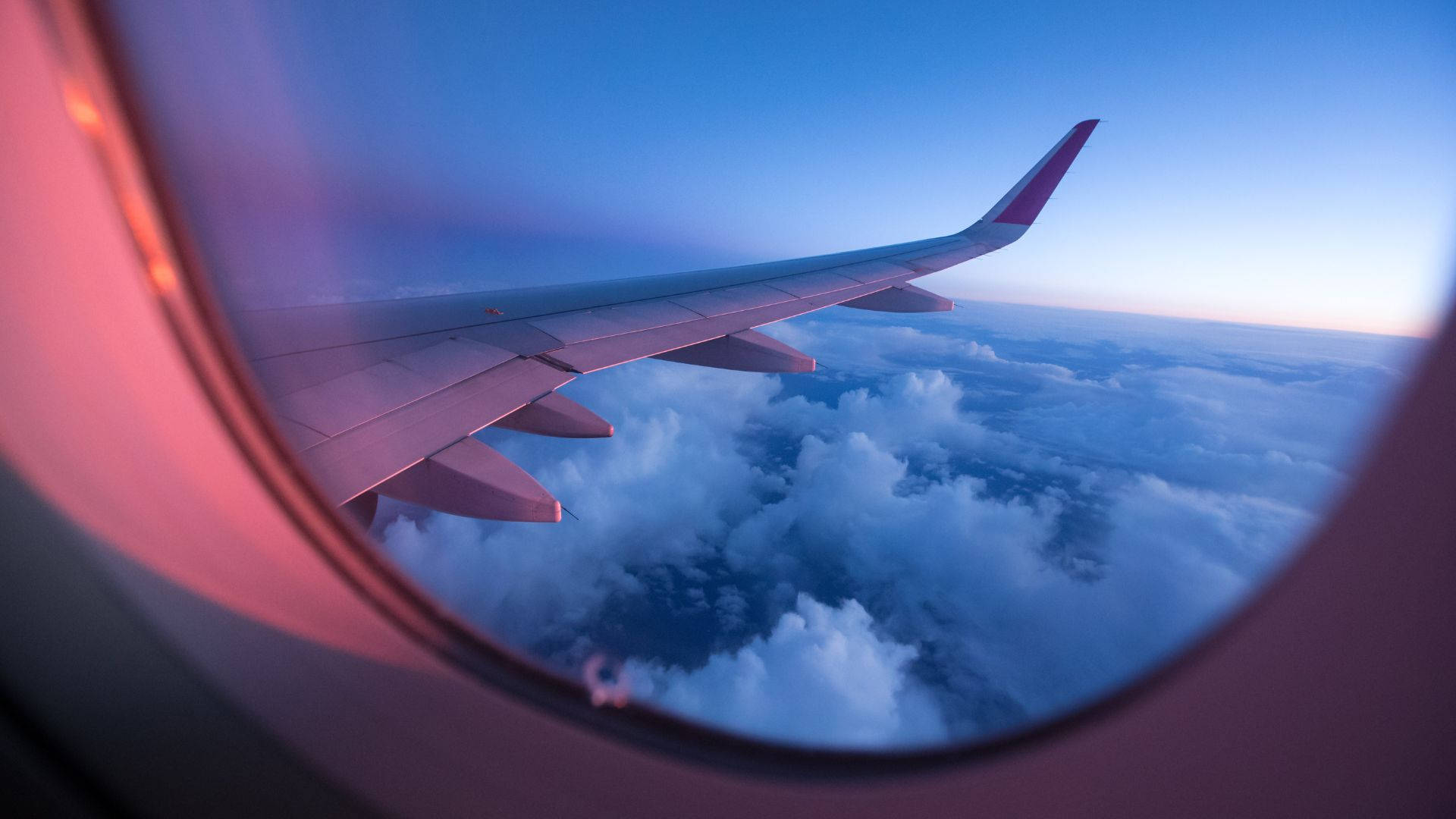 天空景色 桌布 壁紙 — 透過飛機窗戶在黃昏時分看到的飛機機翼，粉紅色和藍色的天空在蓬鬆的雲層之上。
