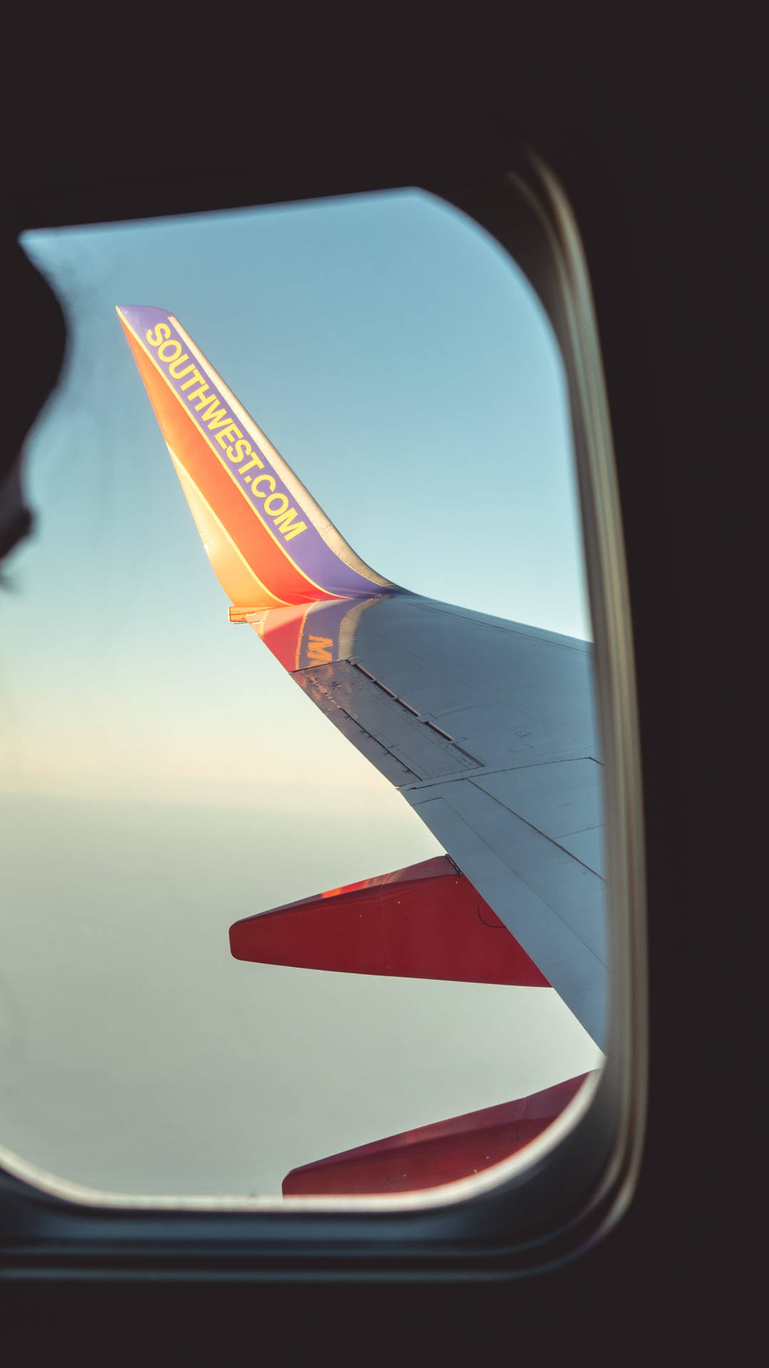 飛機 桌布 壁紙 — 透過飛機窗戶看到西南航空的機翼，背景是日出時清澈的藍天。