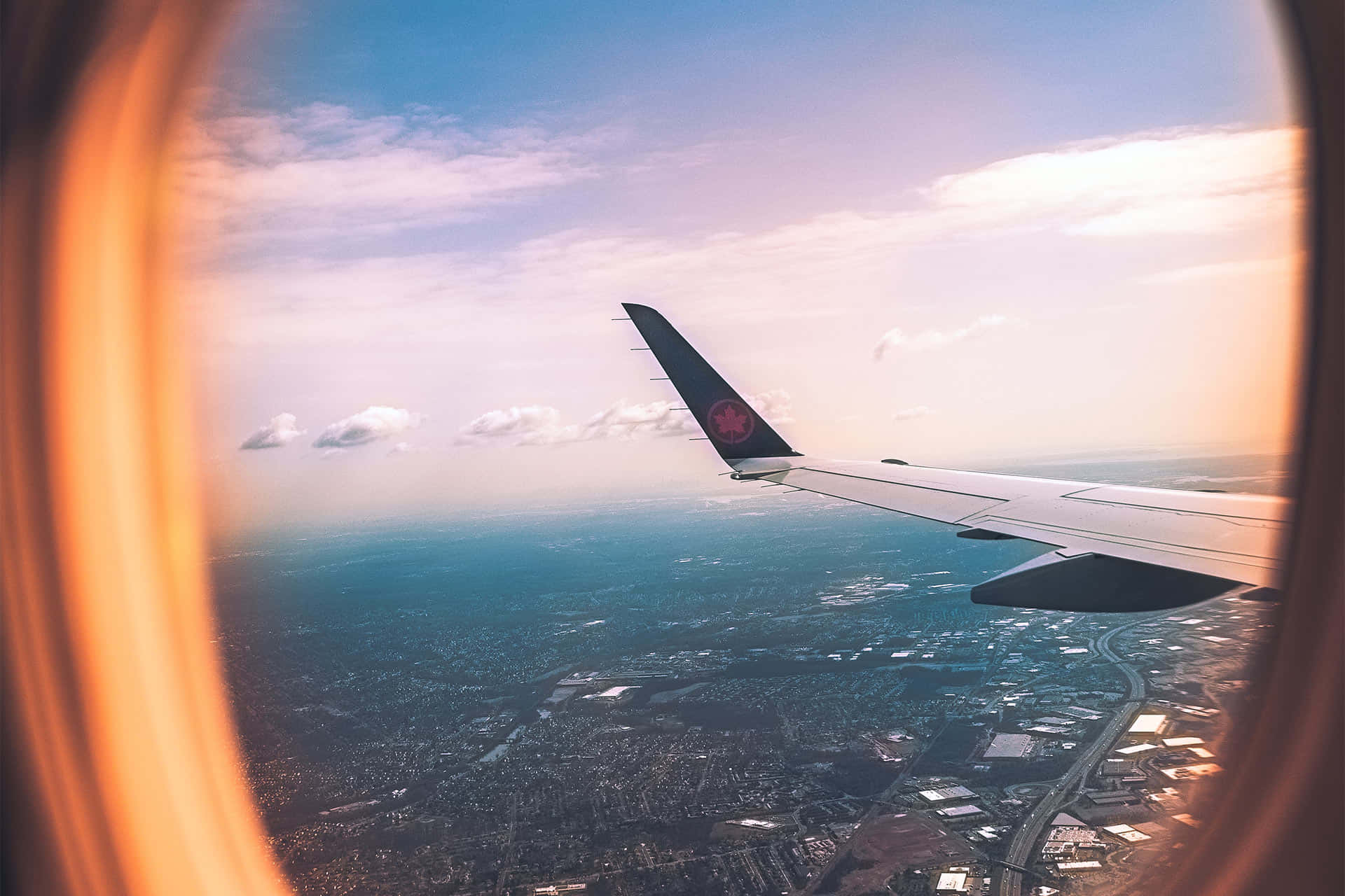 Travel Desktop 桌布 壁紙 — 透過飛機窗戶觀看的飛機機翼，在日落時分，金色光線框住場景，下方是廣闊的城市景觀。