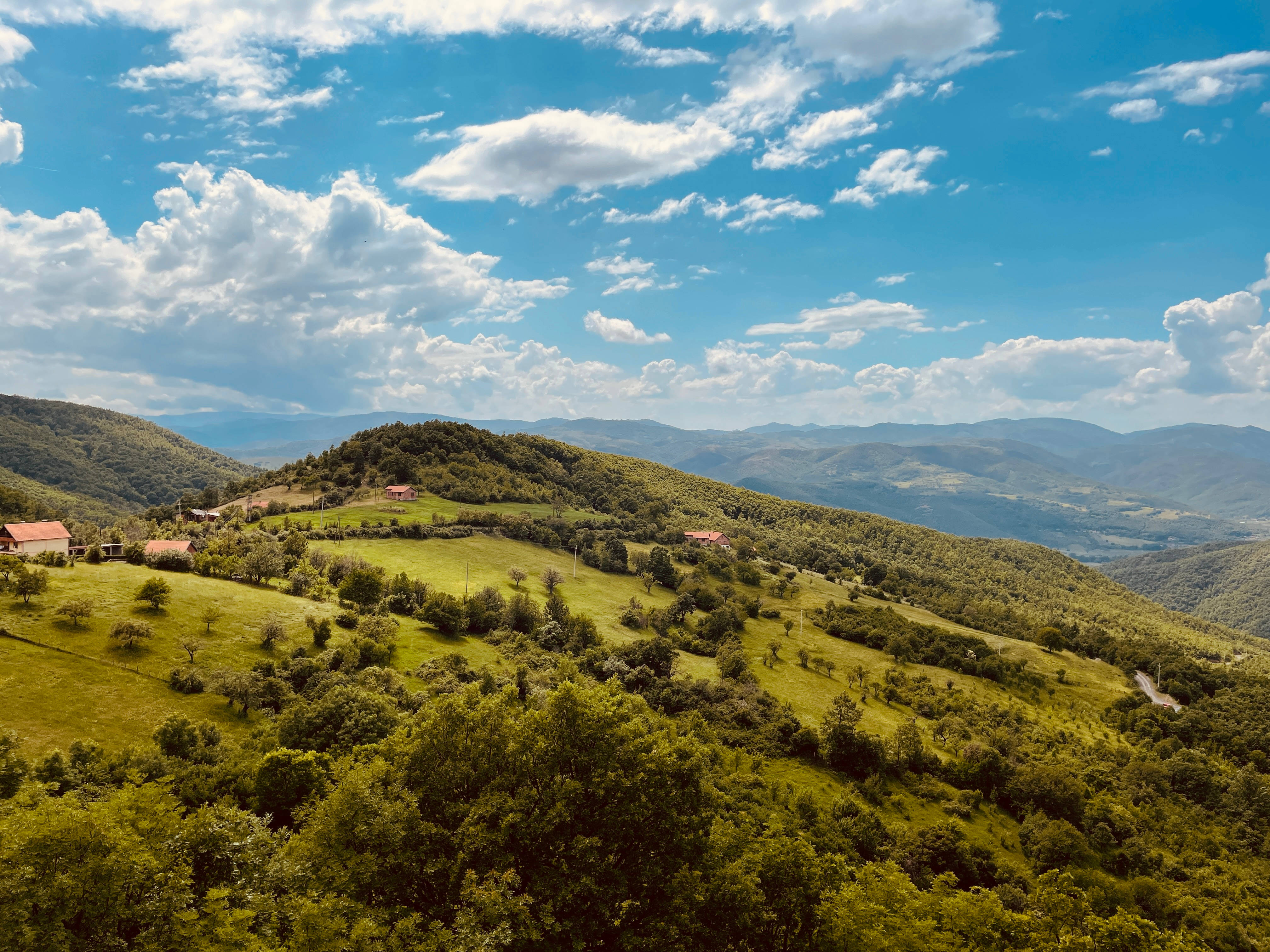 Kosovo 桌布 壁紙 — 連綿起伏的綠色山丘點綴著紅頂房屋，上方是明亮的藍天和蓬鬆的白雲。
