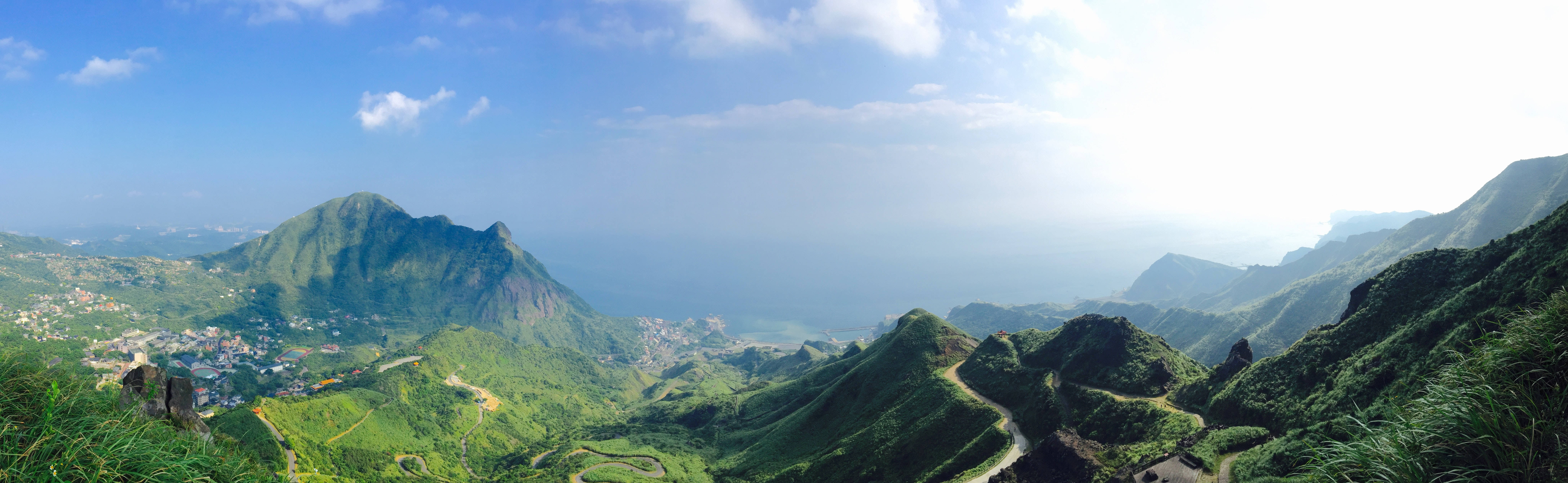 翠綠山脈、蜿蜒的道路和沿海村莊，在明亮的藍天下。 — 4K雙螢幕壁紙 壁紙