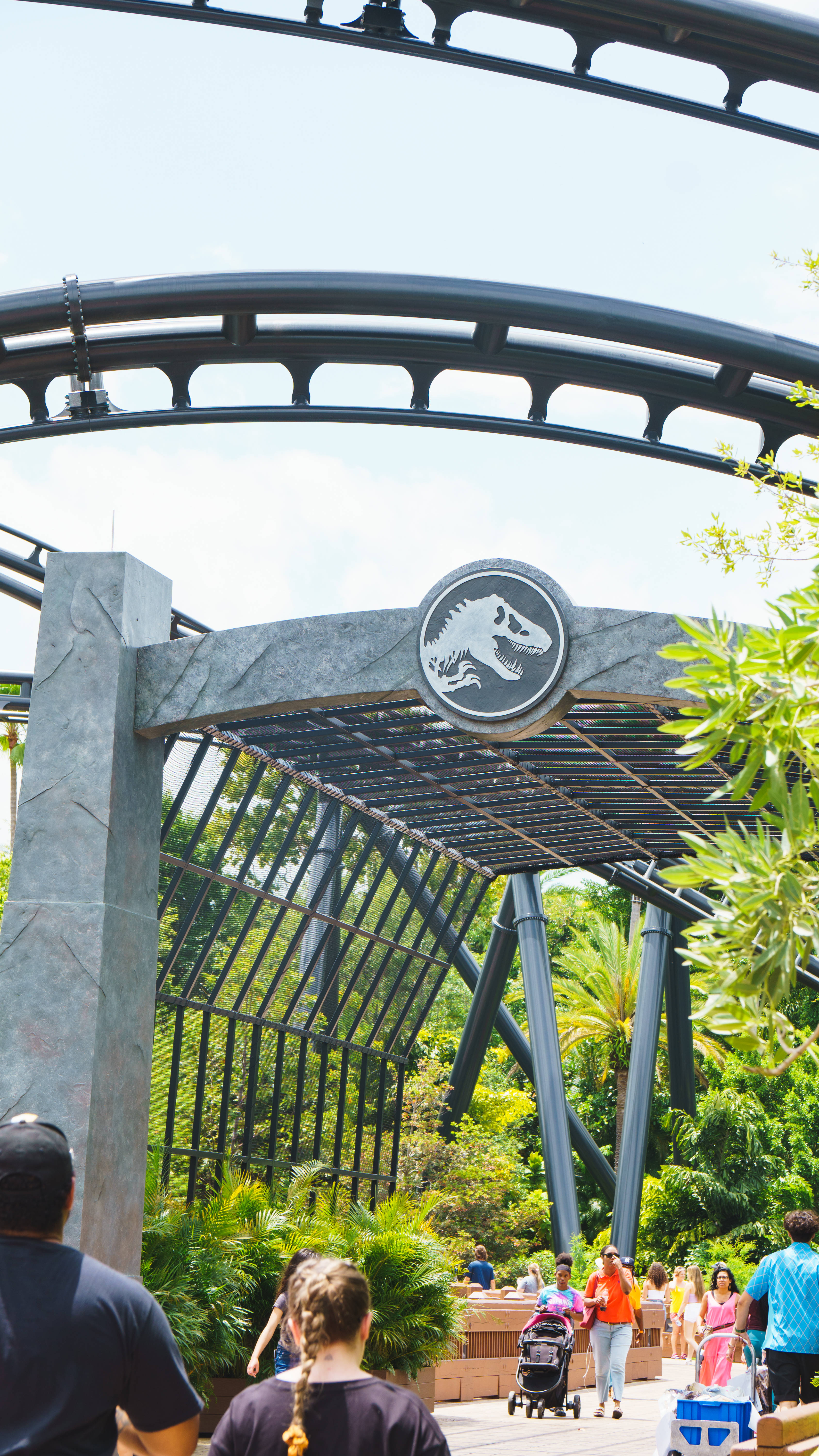 World 桌布 壁紙 — 遊客在侏羅紀公園主題的雲霄飛車入口拱門下行走，拱門上有茂密的綠植和恐龍標誌。