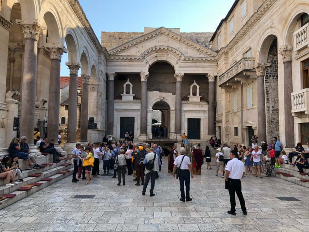 Croatia 桌布 壁紙 — 遊客在晴朗的藍天下探索一個陽光普照的古老石頭庭院，庭院內有宏偉的柱子和拱形步道。