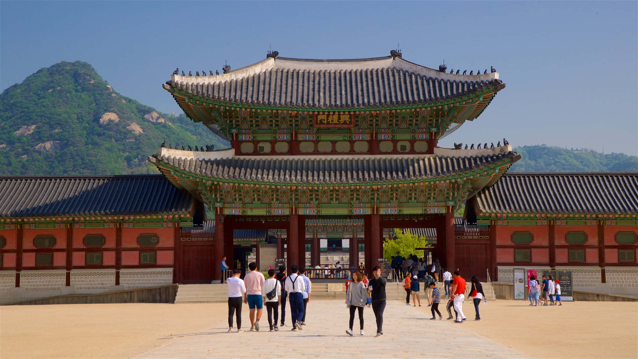 Architecture 桌布 壁紙 — 遊客在晴朗的藍天和綠色山脈背景下，漫步穿過宏偉的傳統韓國宮殿大門。