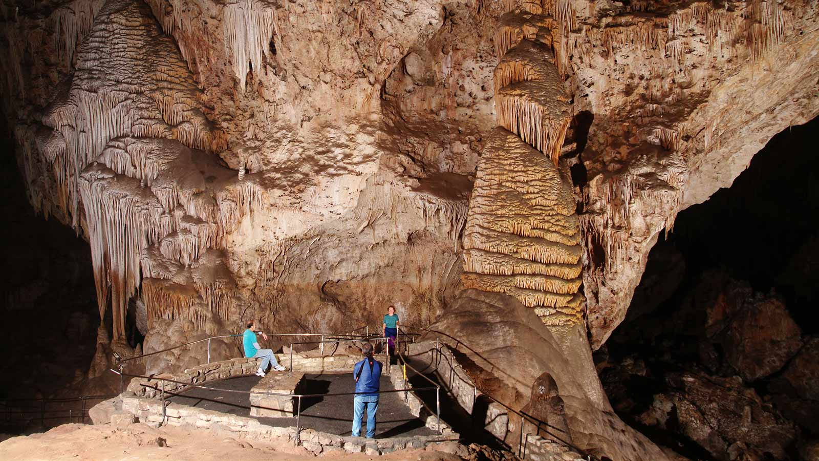Carlsbad Caverns 桌布 壁紙 — 遊客在溫暖的人造燈光下探索一個巨大的石灰岩洞穴，內有高聳的鐘乳石和石筍。