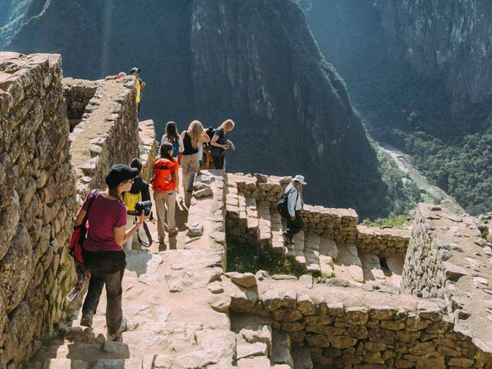 Hiking 桌布 壁紙 — 遊客在燦爛陽光下攀登馬丘比丘的古老石階梯，下方是陡峭的山崖和鬱鬱蔥蔥的山谷。
