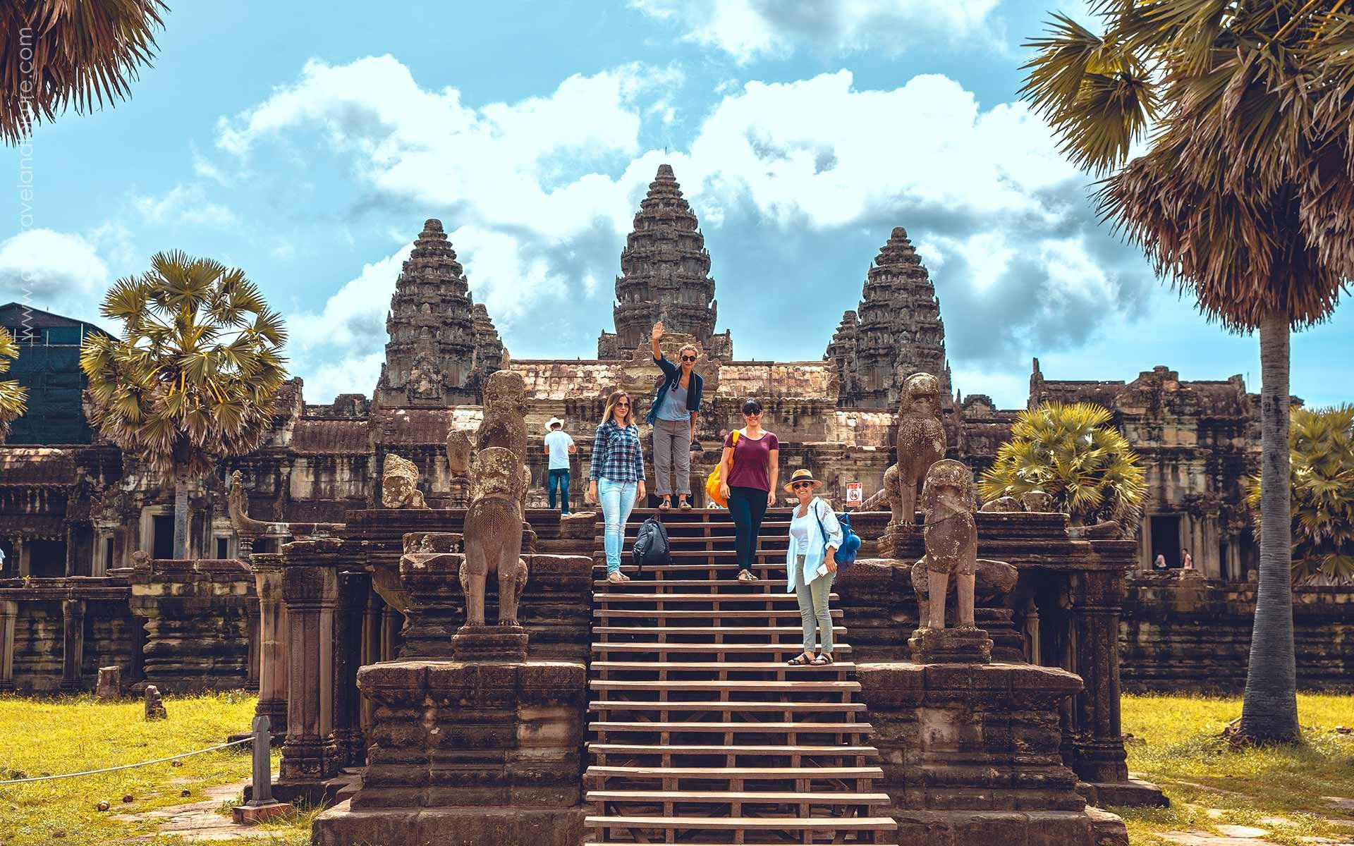 Cambodia 桌布 壁紙 — 遊客在藍天白雲下，於雄偉的吳哥窟寺廟前，在兩旁有獅子雕像的石階上擺姿勢。