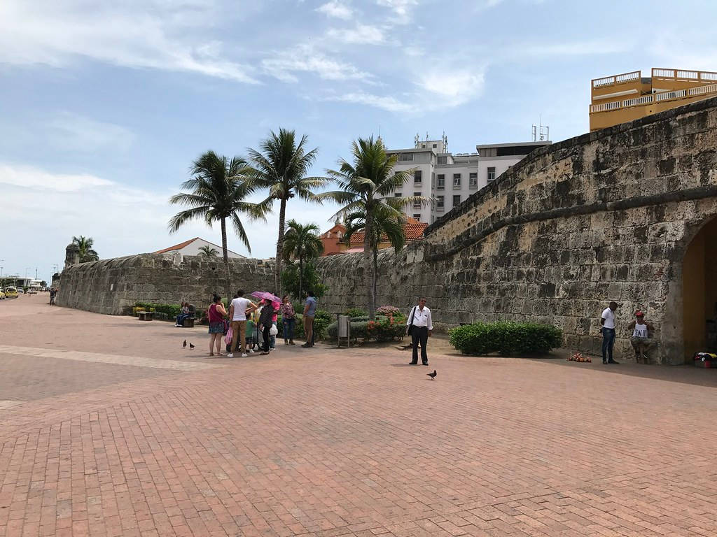 Colombia 桌布 壁紙 — 遊客在部分多雲的天空下，沿著陽光照耀的磚砌廣場，在古老石牆和棕櫚樹旁邊漫步。