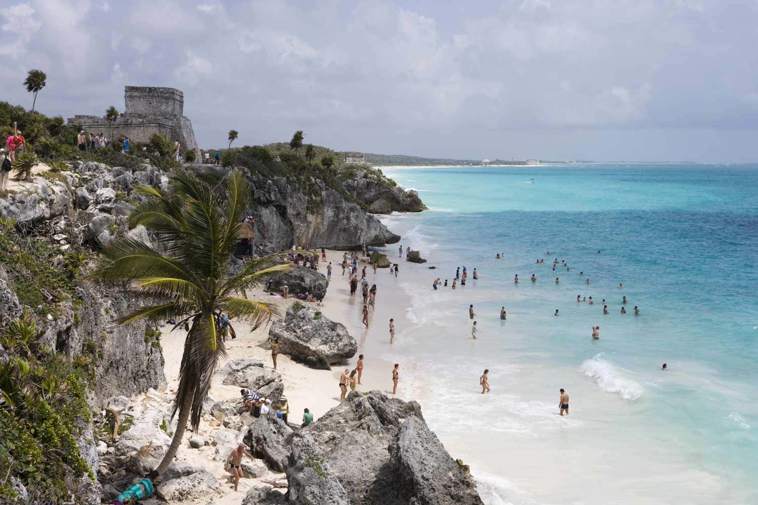 Mexican Beach 桌布 壁紙 — 遊客在陰天之下，於懸崖海灘上享受碧綠海水和白色沙灘，旁邊有古代遺跡和棕櫚樹。