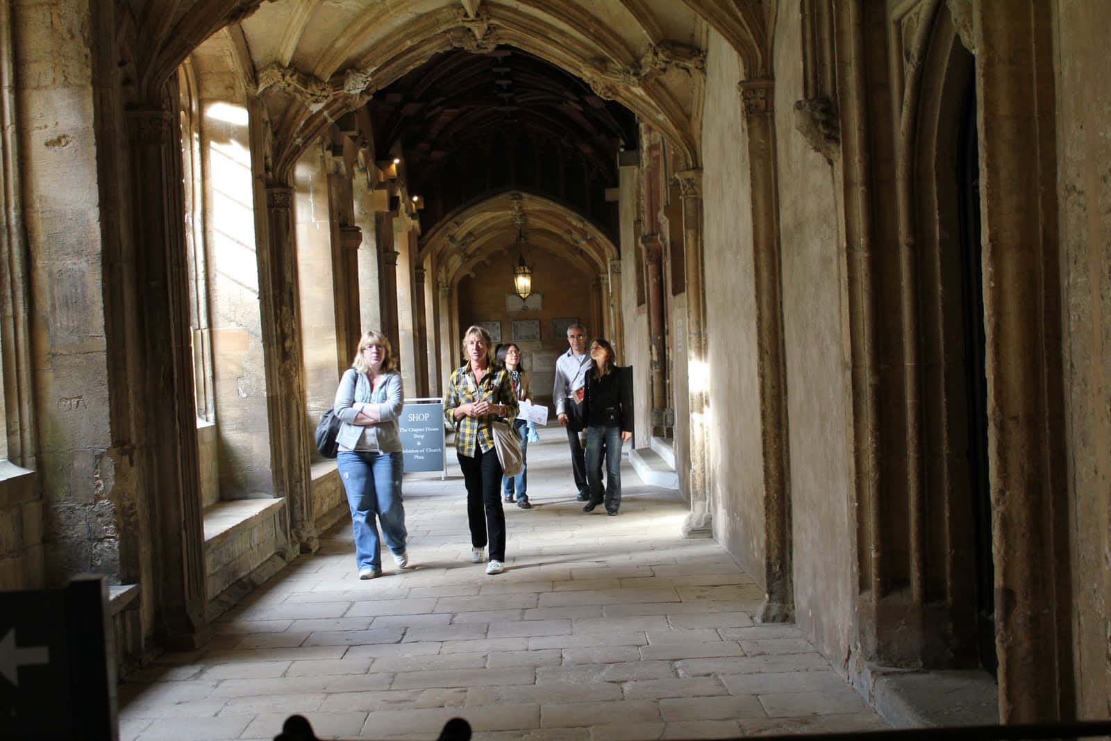 The Hogwarts Charms Corridor 桌布 壁紙 — 遊客在陽光普照的石頭迴廊中漫步，拱形走道和歷史柱廊相伴。