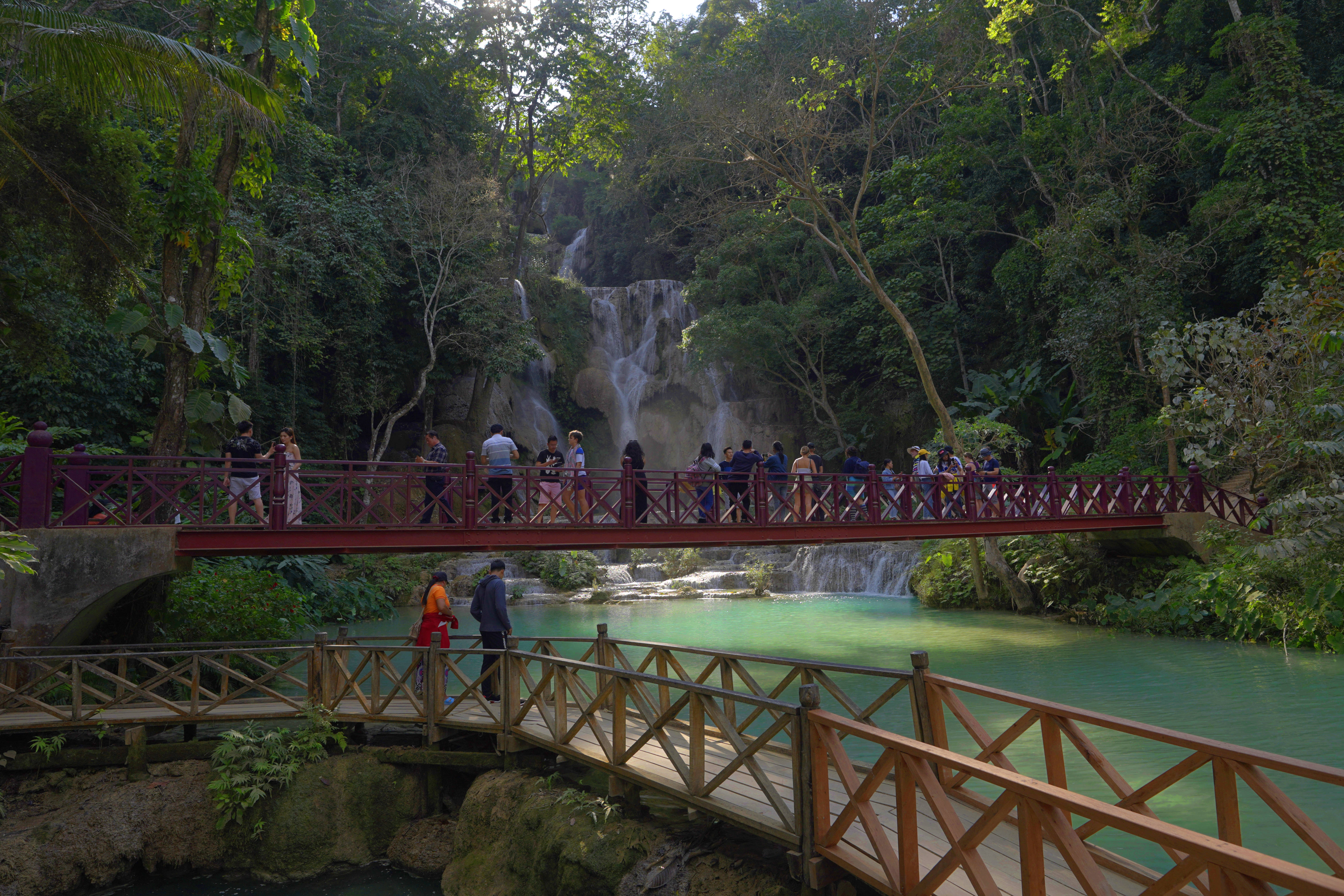 Laos 桌布 壁紙 — 遊客漫步在橫跨碧綠水面的紅色橋樑上，多層瀑布穿過茂密的綠色叢林傾瀉而下。