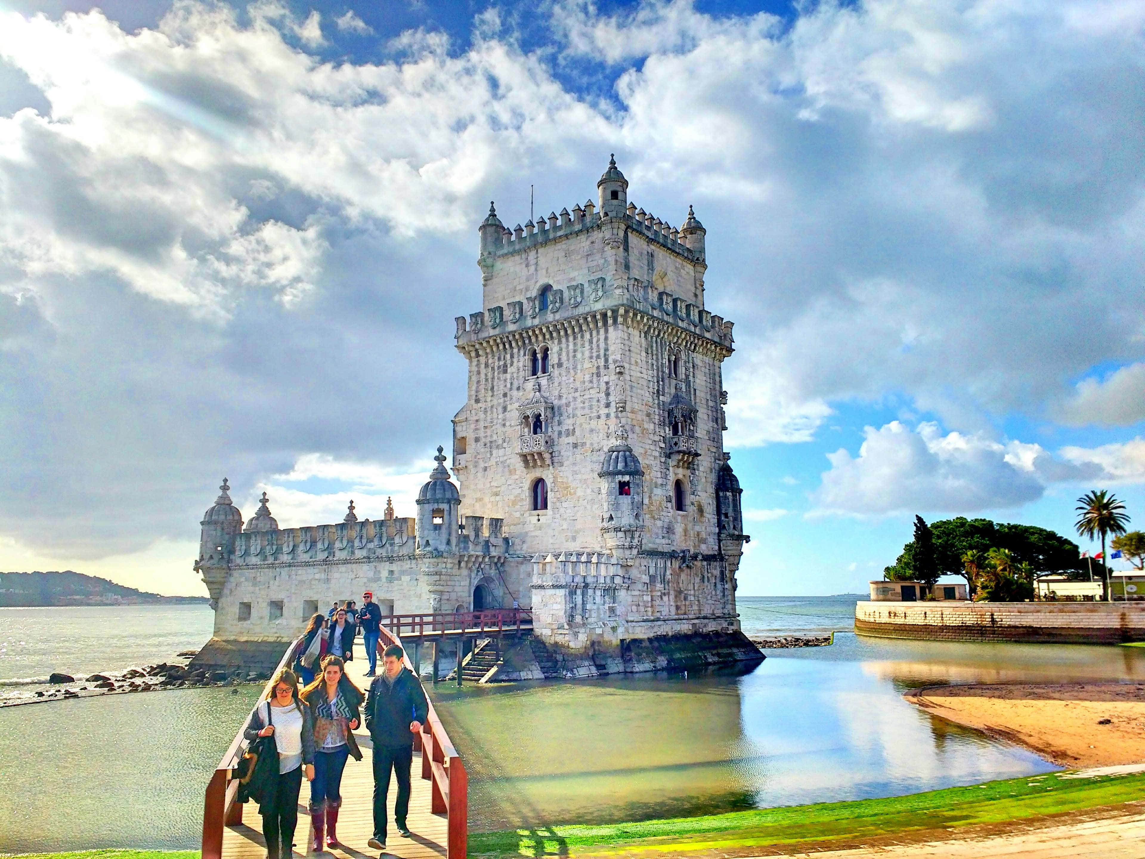 Portugal 桌布 壁紙 — 遊客漫步穿過一座紅色橋樑，前往歷史悠久的貝倫塔，該塔矗立在平靜的水面上，天空部分多雲。