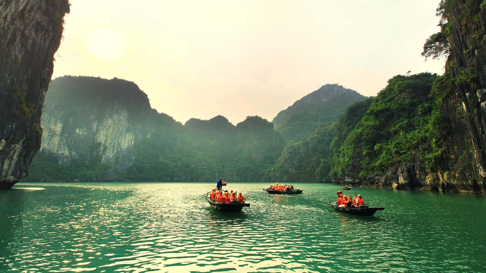 Vietnam 桌布 壁紙 — 遊客穿著橙色救生衣，在黃金時段划著小船穿過寧靜的翡翠色潟湖，兩旁是高聳的叢林覆蓋的石灰岩懸崖。
