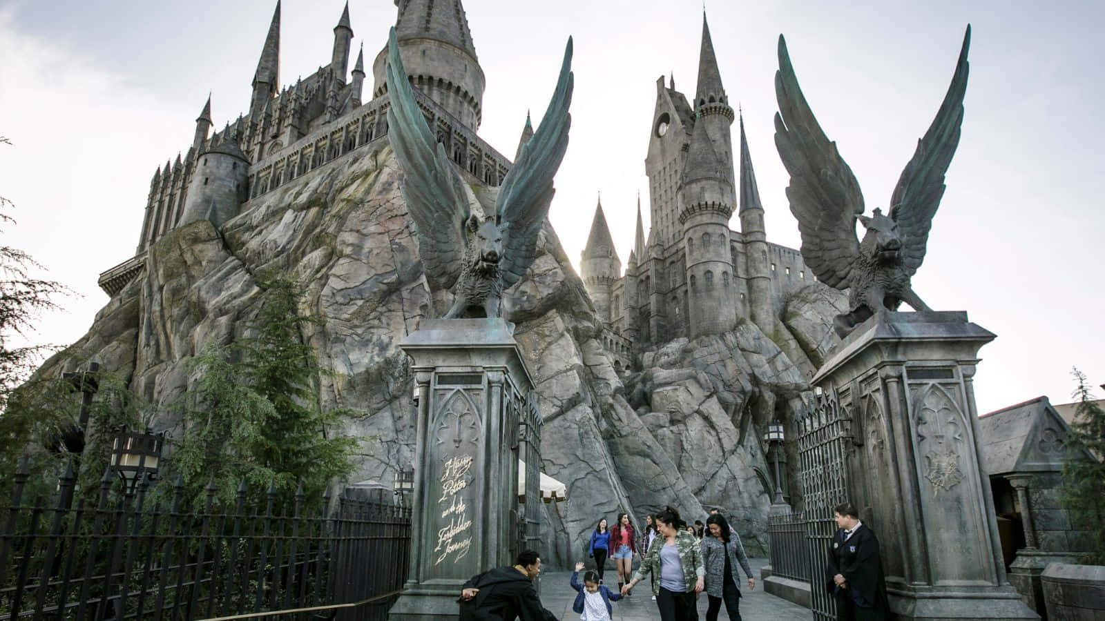 Wizarding World 桌布 壁紙 — 遊客穿過華麗的大門，兩側有帶翅膀的雕像，走向坐落在岩石懸崖上的高聳哥德式霍格華茲城堡。