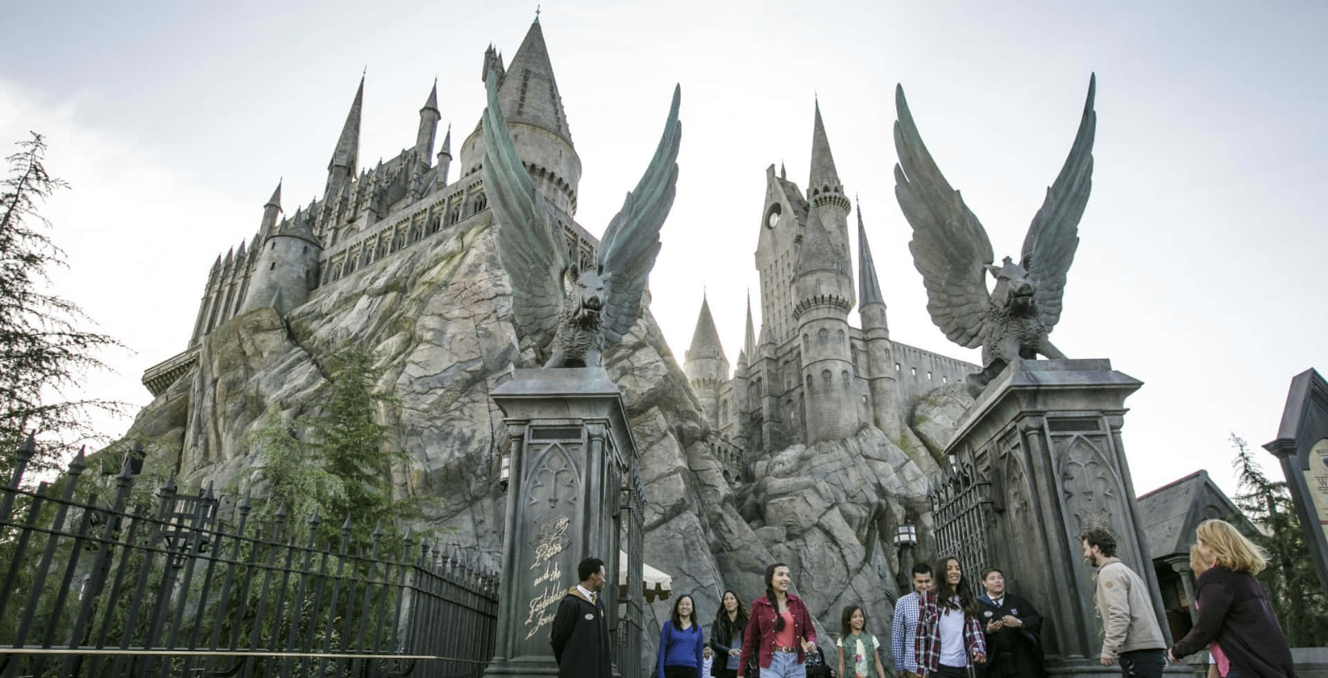 Wizarding World 桌布 壁紙 — 遊客站在宏偉的霍格華茲城堡前，城堡擁有高聳的尖塔和帶翅膀的石像鬼雕像，天空多雲。