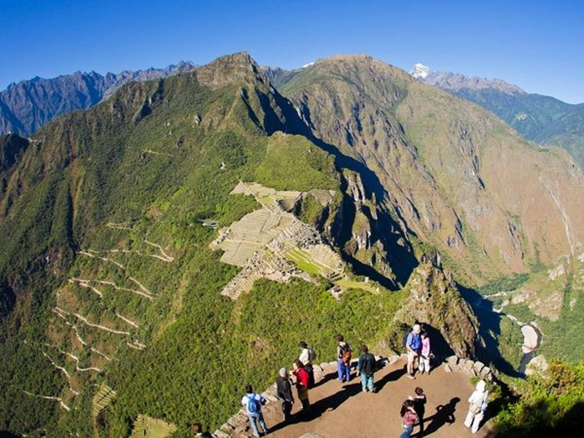 Hiking 桌布 壁紙 — 遊客站在懸崖上俯瞰馬丘比丘的古老石頭遺址，這些遺址坐落在鬱鬱蔥蔥的綠色山脈中，天空清澈湛藍。