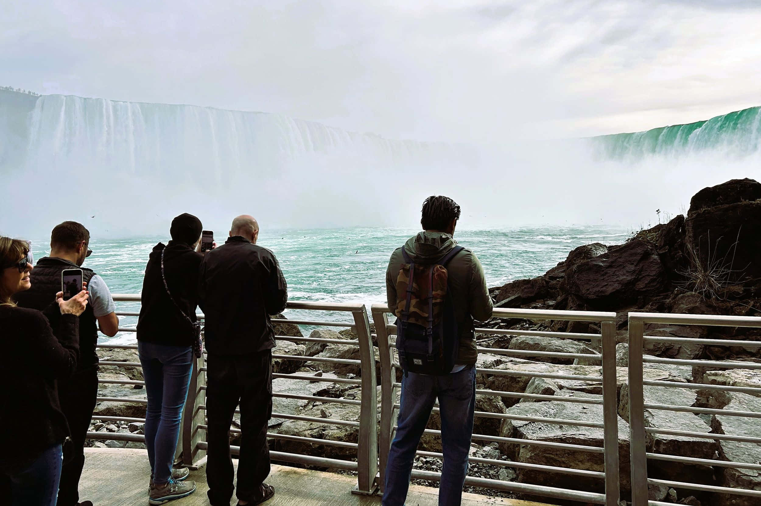 Niagara Falls 桌布 壁紙 — 遊客站在欄杆旁，在陰天拍攝巨大的霧濛濛尼亞加拉瀑布。